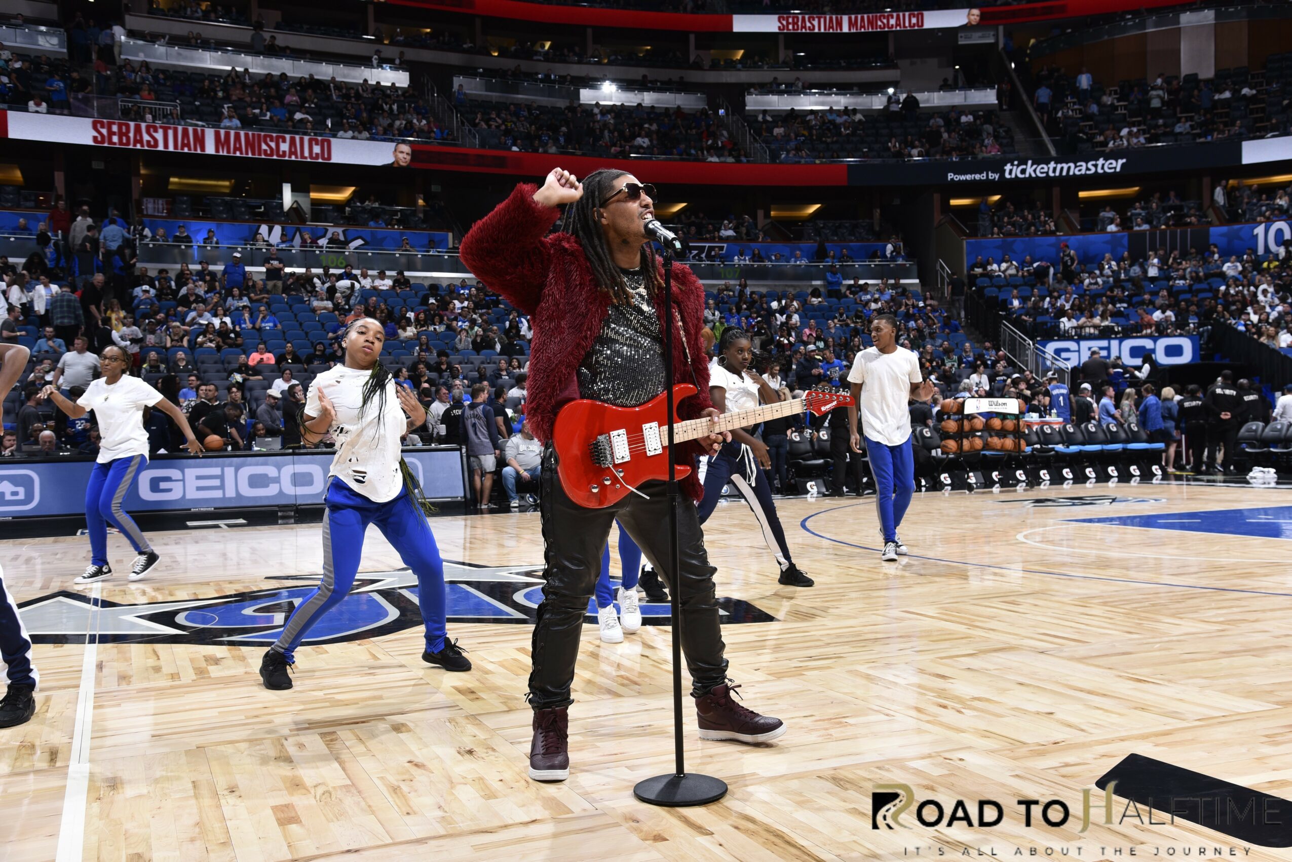 Musician with red guitar performing on basketball court, people dancing in background, arena setting, stage lights, audience seats.