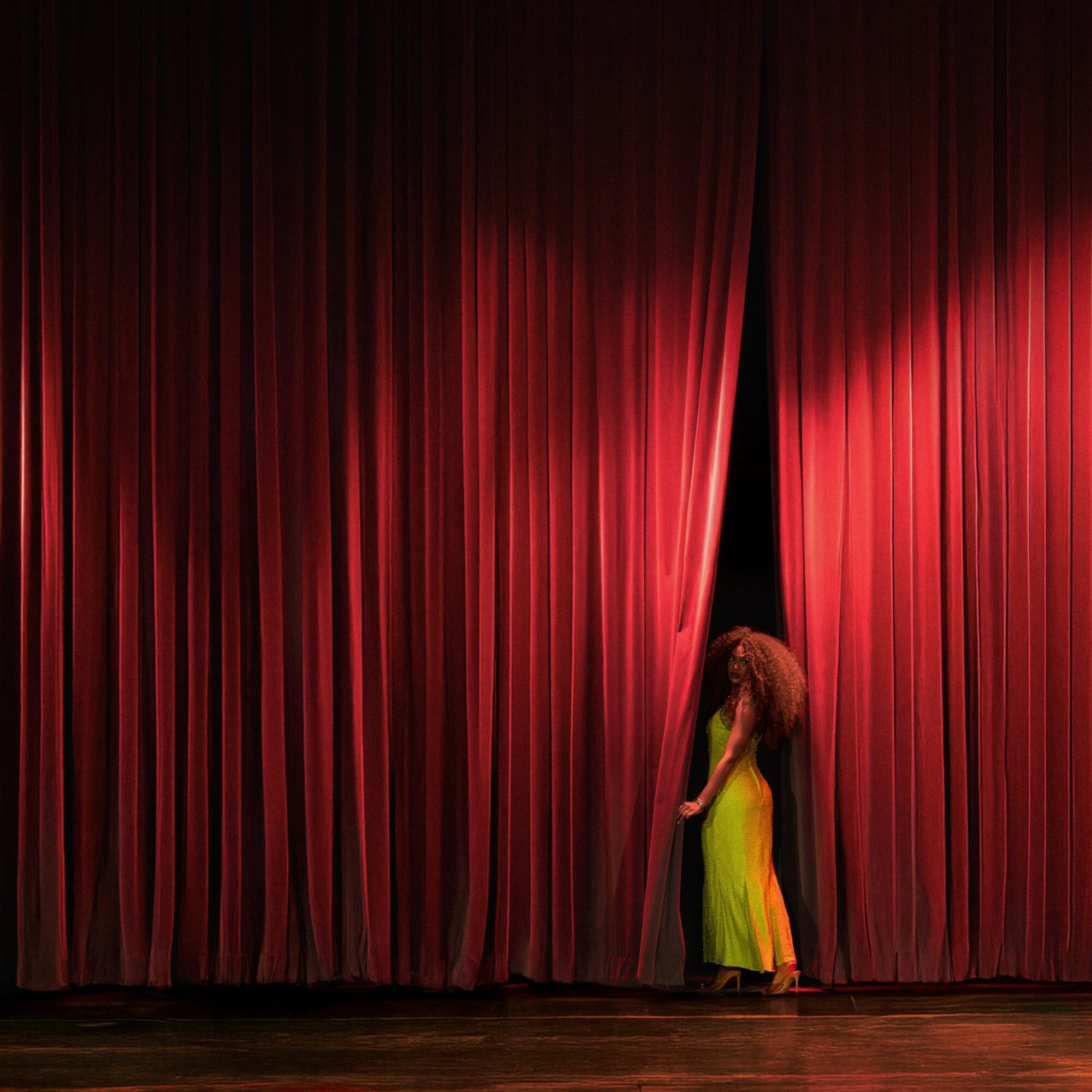 Person in a yellow dress stands behind red stage curtains, partially opening them, on a wooden stage.
