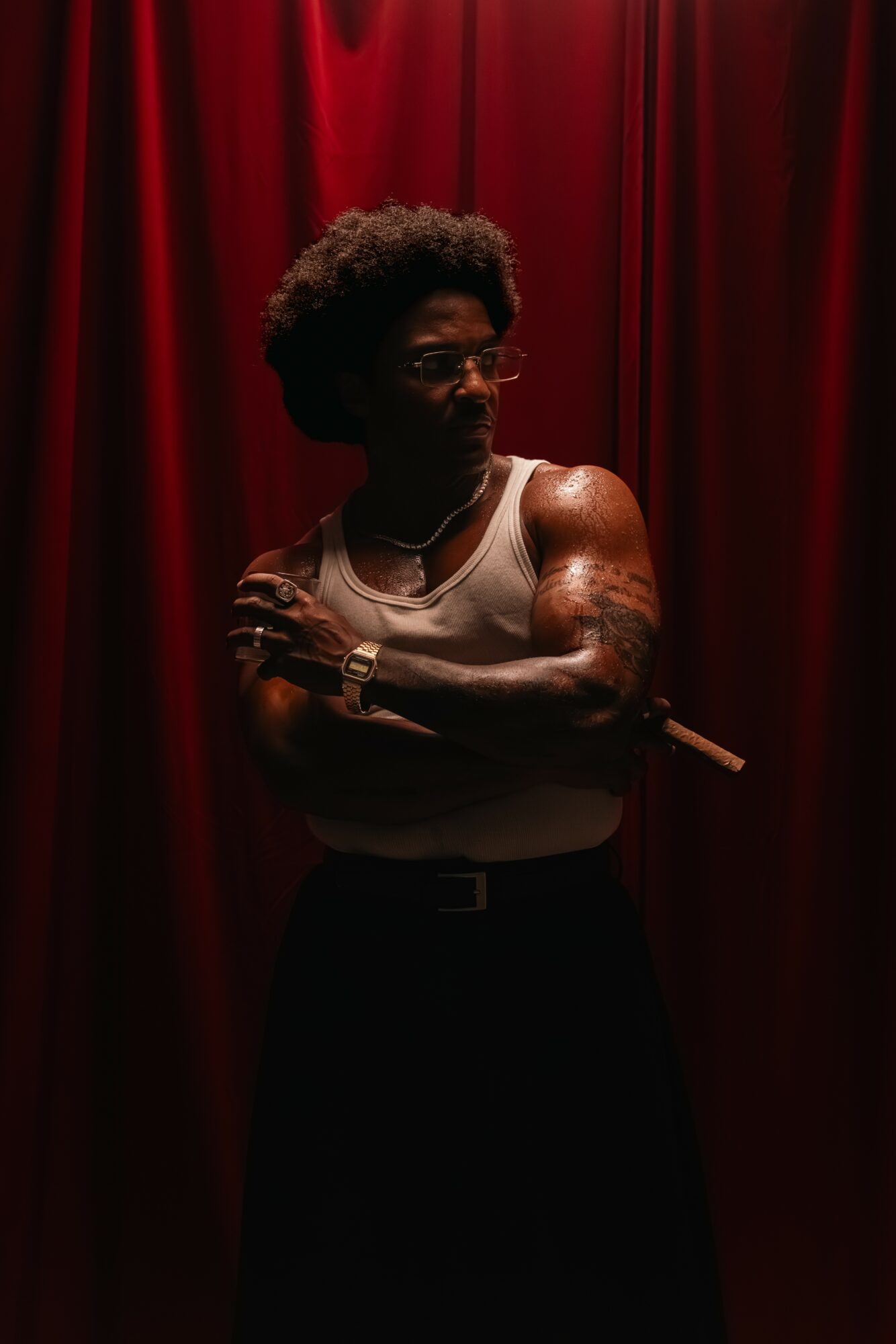 Person with curly hair and glasses stands with arms crossed in front of red curtains, wearing a sleeveless top and dark pants.