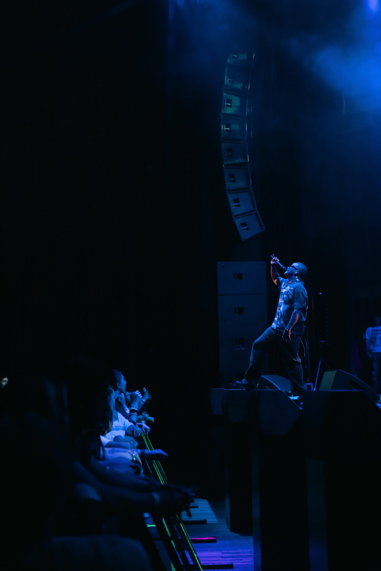 Performer on stage with blue lighting, microphone, and speakers, audience seated in front, dark background.