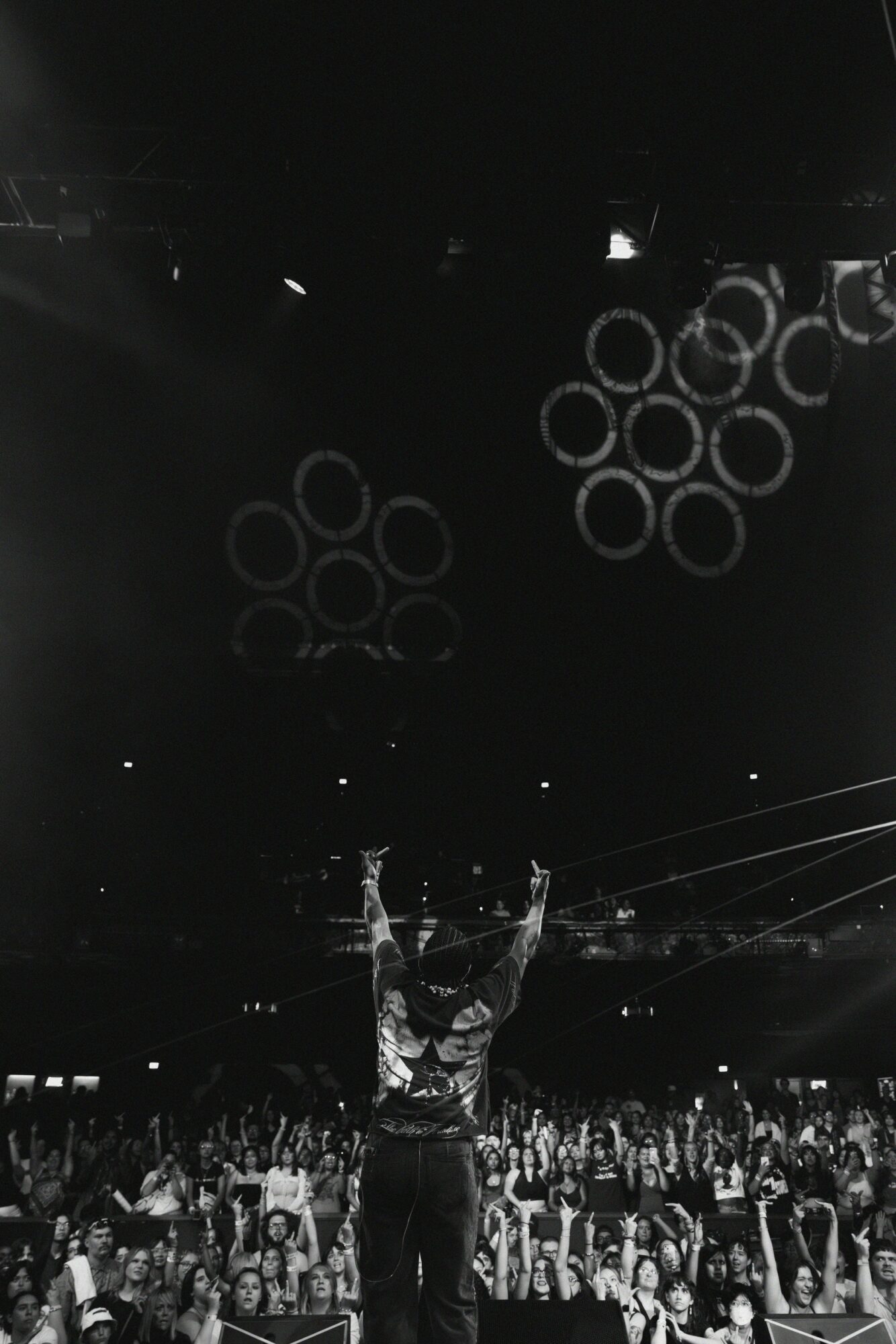 Person on stage with arms raised in front of a large audience, stage lights, and circular light patterns.