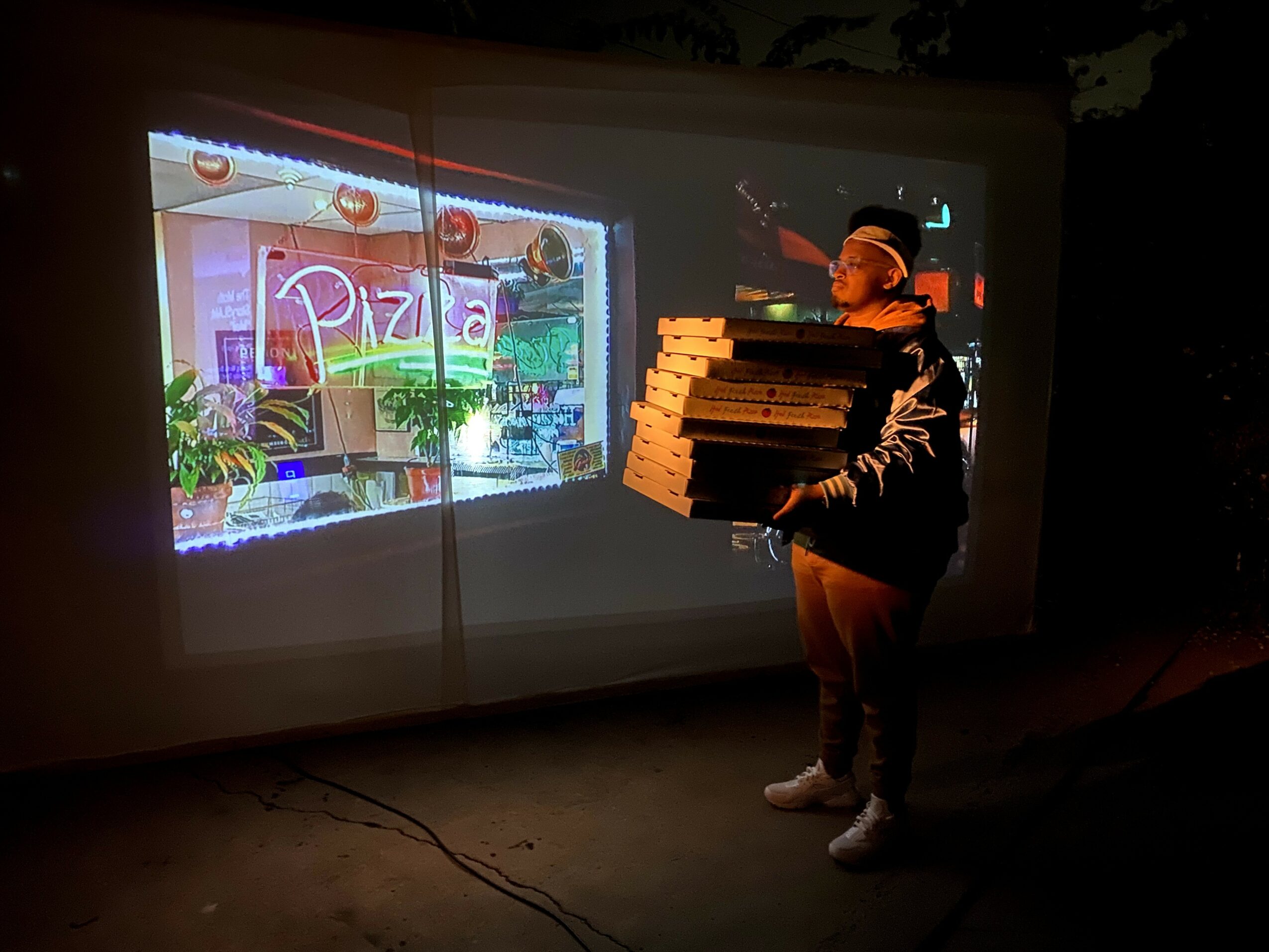 Person holding multiple pizza boxes standing in front of a projected pizza shop window display.