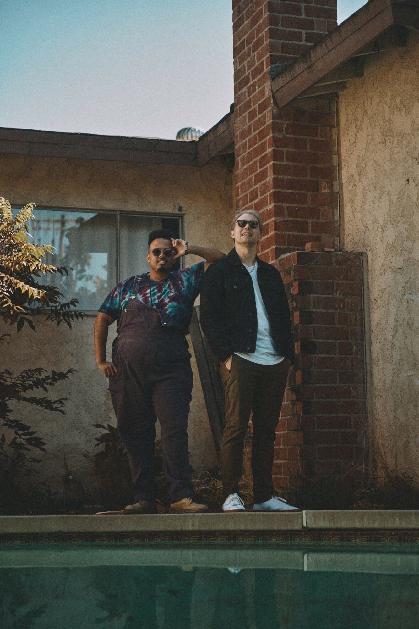 Two men stand outdoors near a brick wall and a building, one wearing sunglasses and a patterned shirt, the other in a black jacket and white shirt, with a pool in the foreground.