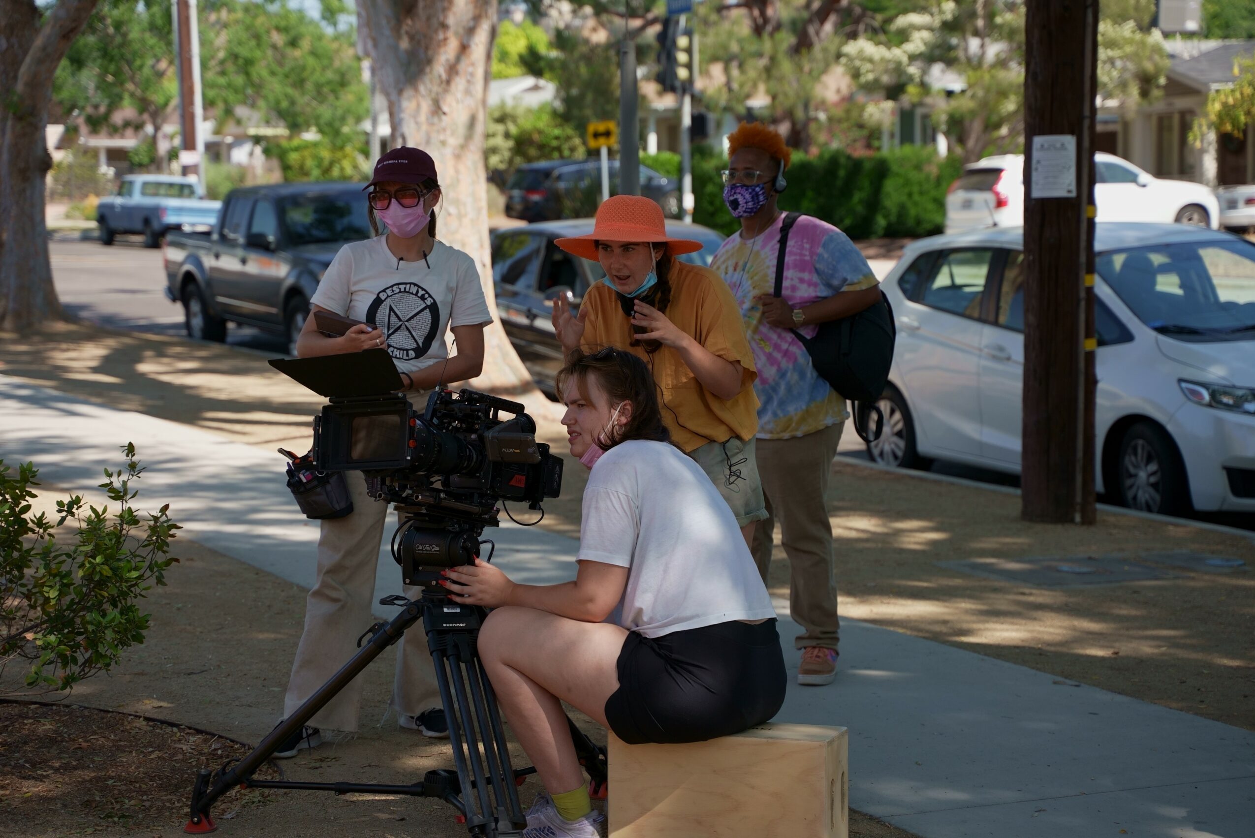 Four people, one sitting on a wooden block with a camera, three standing nearby, on a sidewalk with parked cars and trees.