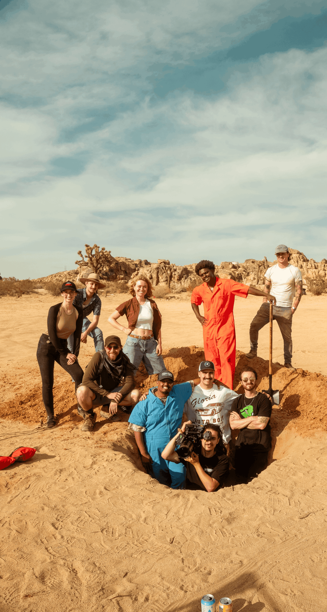 Group of nine people outdoors in a sandy area with a blue sky and clouds, some standing and some sitting or kneeling.