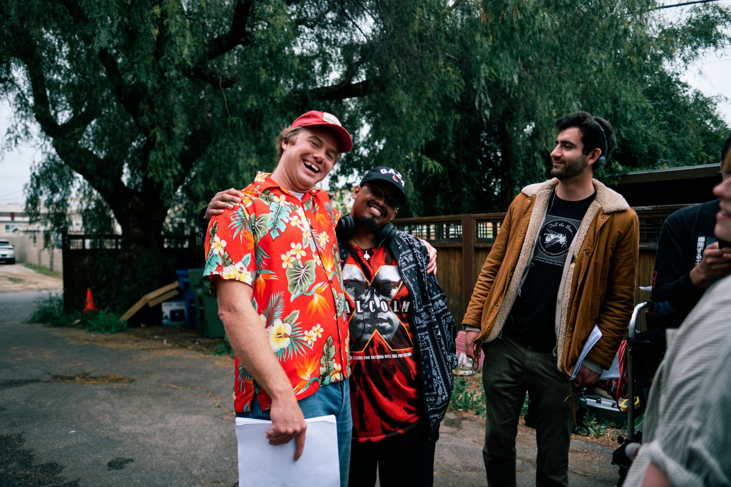 Four people outdoors, smiling and talking, with trees and a parking lot in the background.