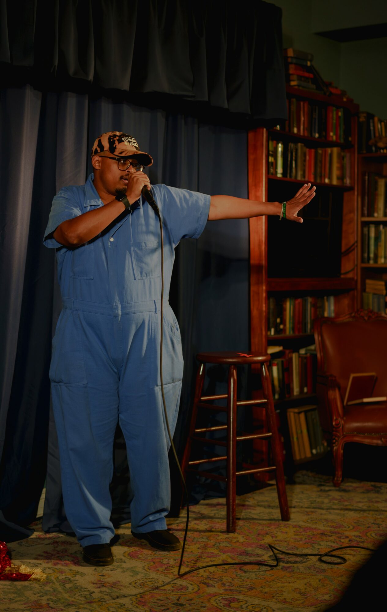 Person in blue jumpsuit holding microphone, gesturing with arm extended, standing on stage with bookshelves in background.