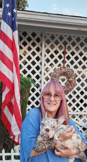Woman with pink hair and glasses holding a dog, standing outdoors near a house with a white lattice fence and an American flag.