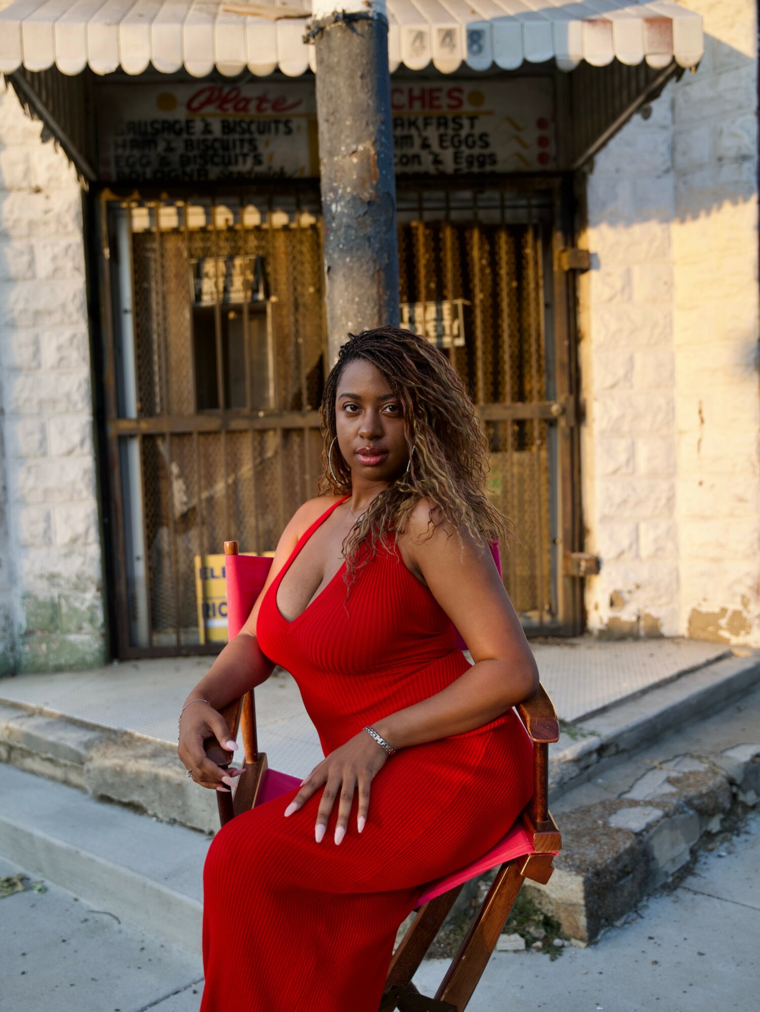 Woman in red dress sitting on a wooden chair outside a building with a wooden gate and brick wall.