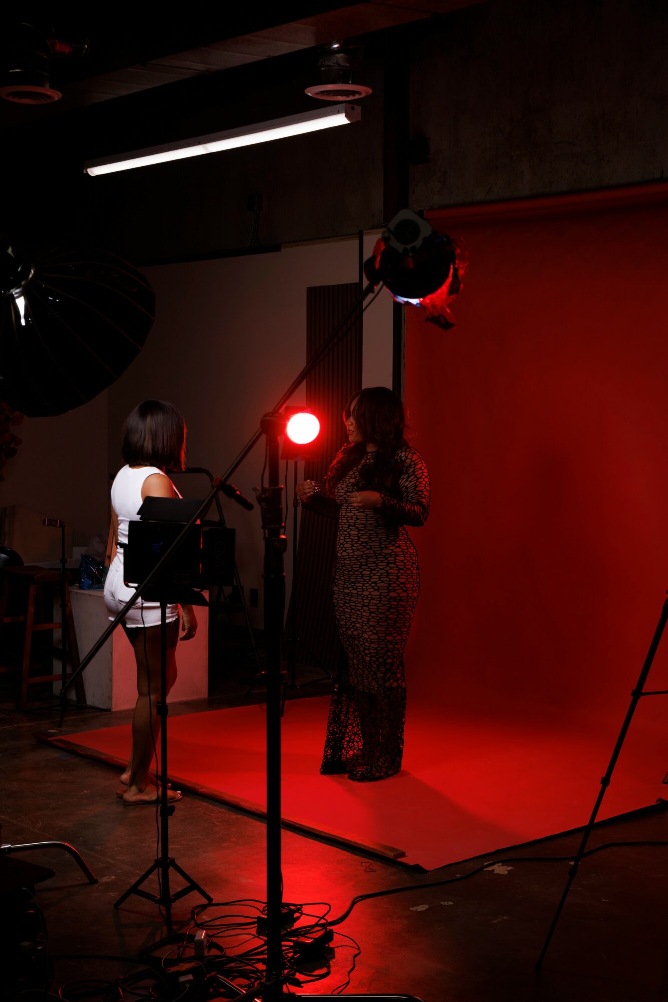 Two women stand in a photography studio with a red backdrop, illuminated by studio lights and equipment.