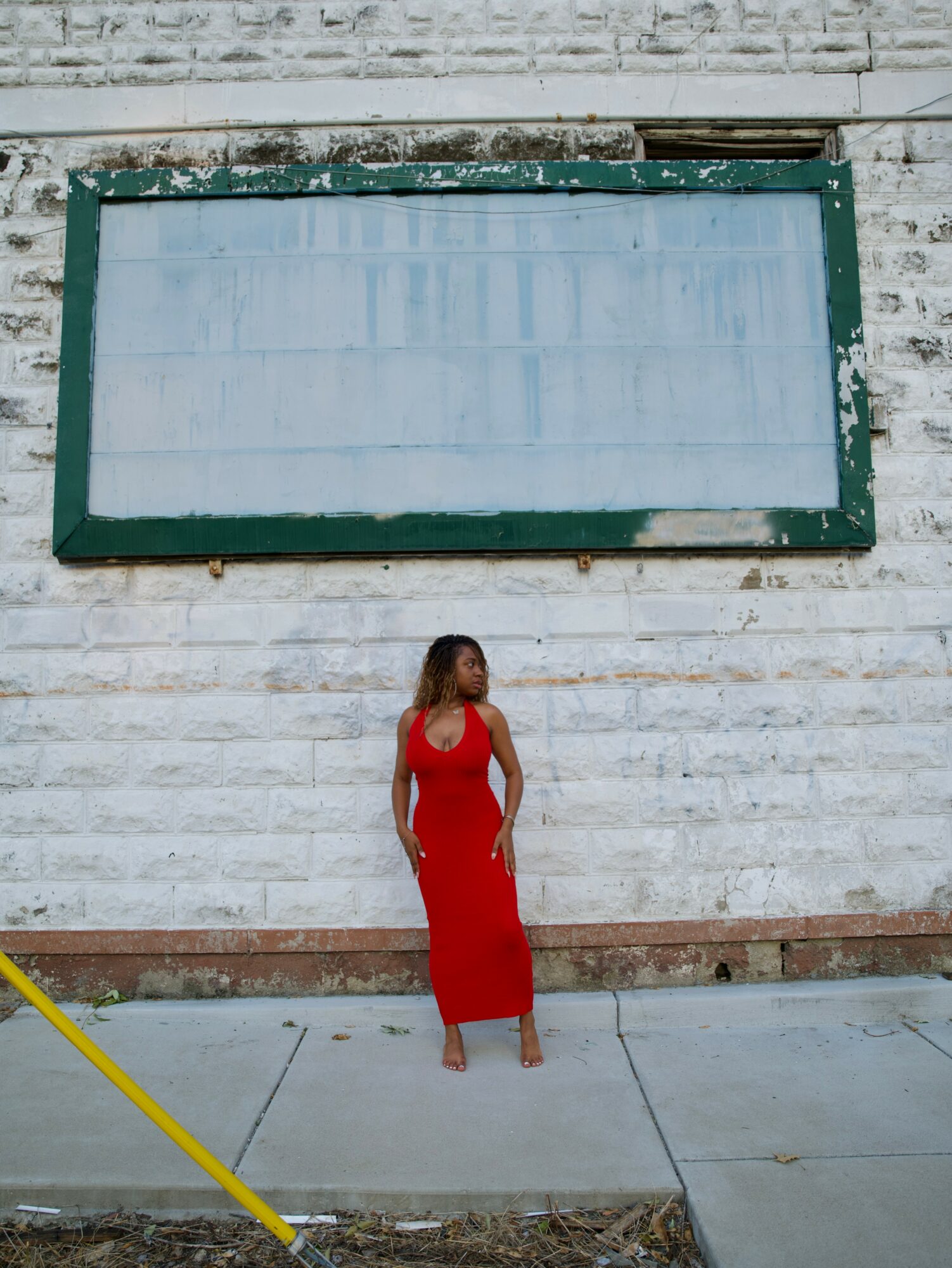 Woman in red dress standing on sidewalk in front of a white brick wall with a large, empty signboard above her.