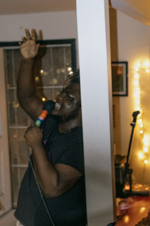 Person singing into a microphone, raising one hand, standing behind a white partition in a dimly lit room.