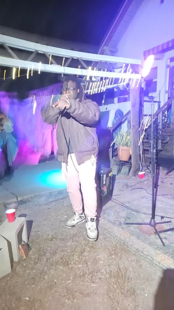 Person standing outdoors at night, holding a microphone, with colorful lights and a railing in the background.