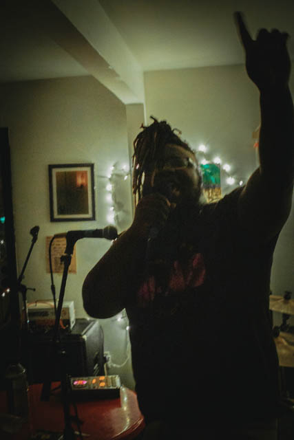 Person taking a mirror selfie with one hand raised, in a dimly lit room with musical equipment and a framed picture.