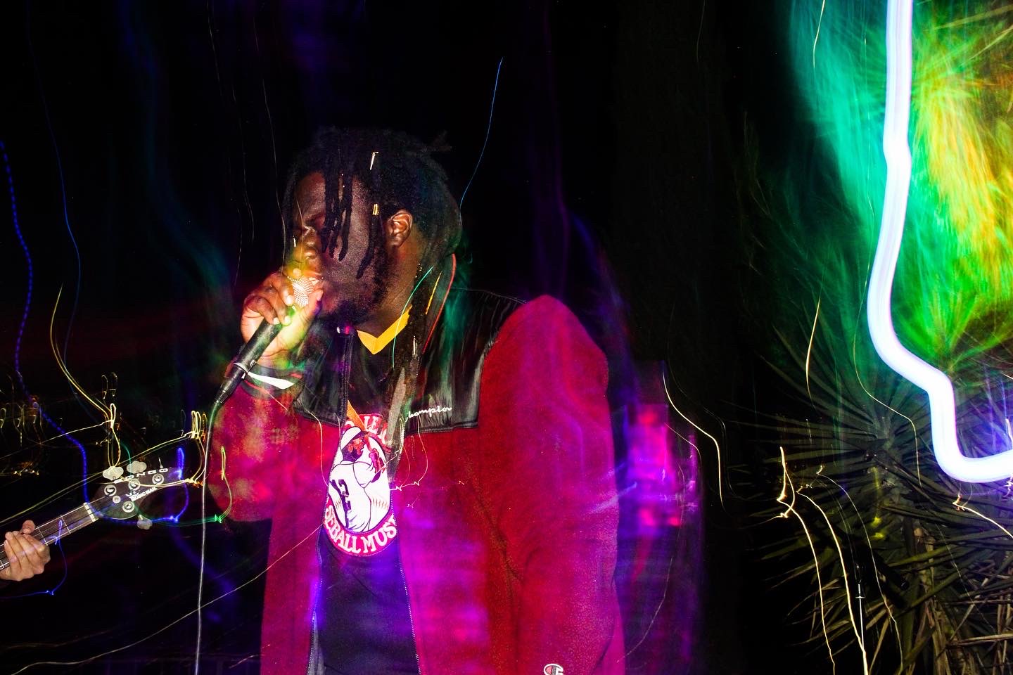Person with dreadlocks singing into a microphone, wearing a red hoodie with a white logo, colorful light streaks around.