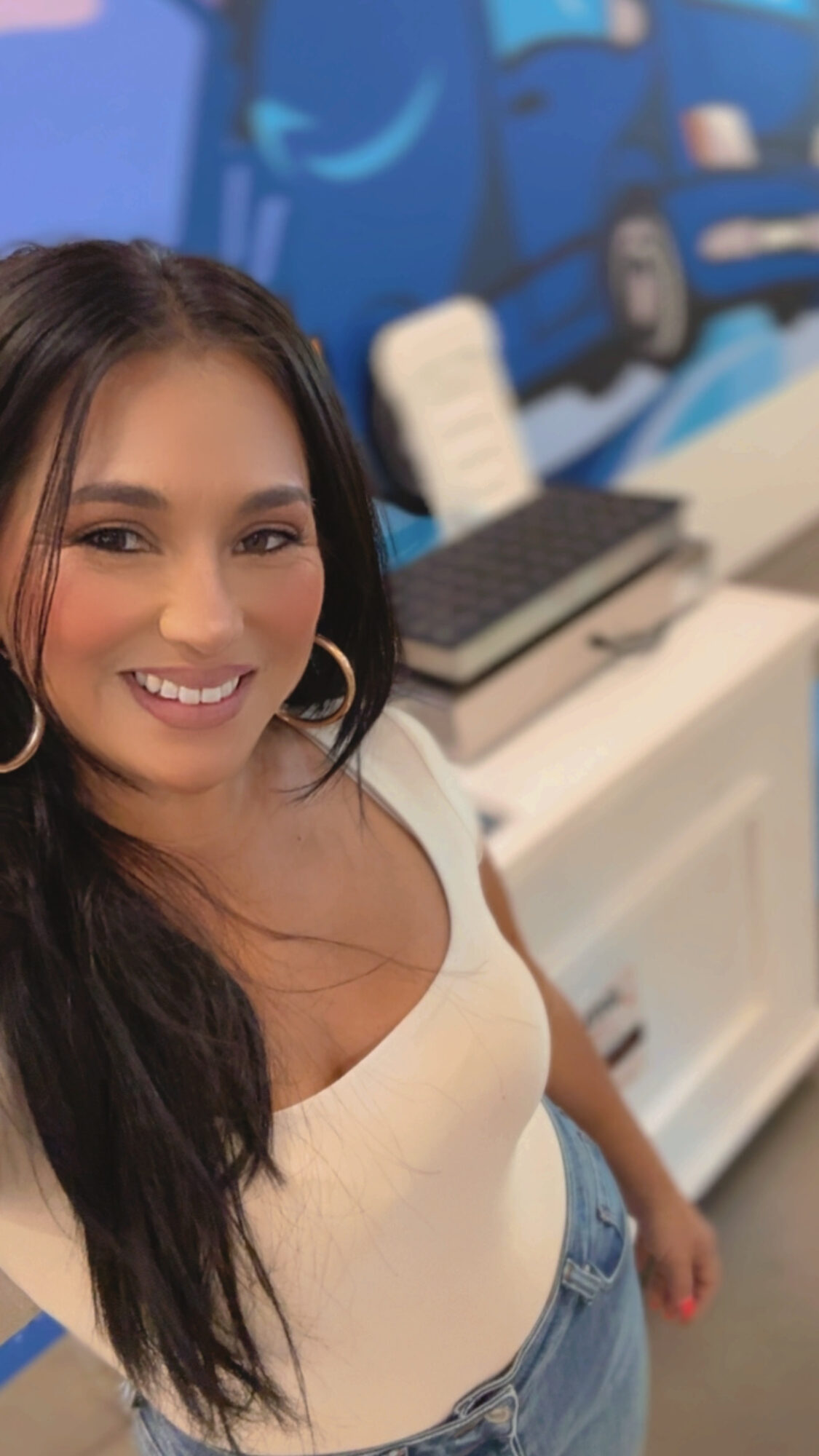 Smiling woman with long dark hair wearing a white top and jeans, standing indoors with a colorful background.