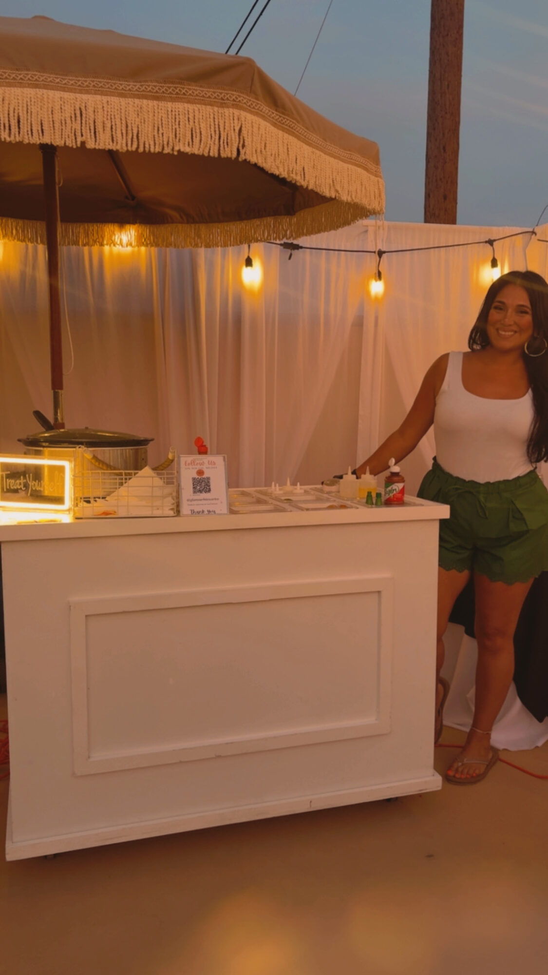 Woman standing at a white outdoor bar with string lights and a canopy, smiling, wearing a white top and green shorts.