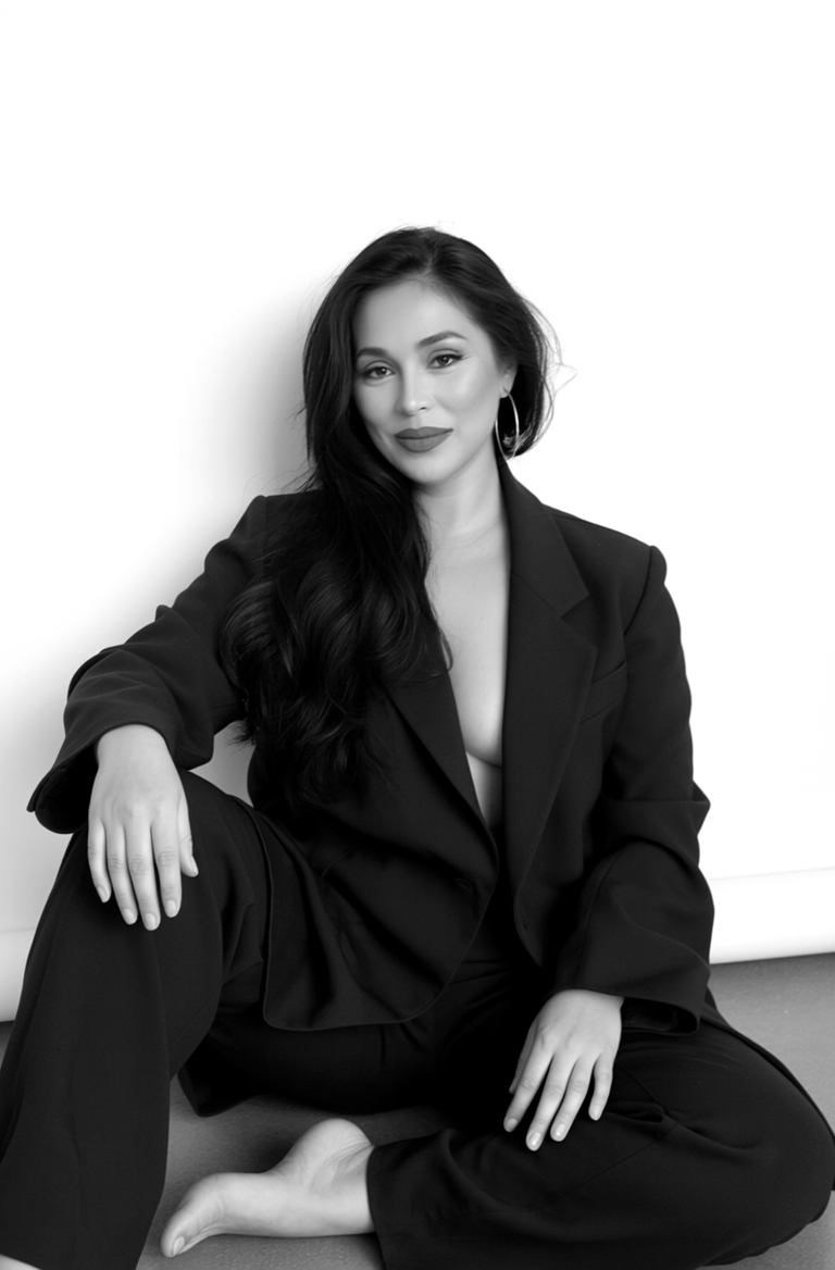 Woman with long dark hair sitting on the floor, wearing a blazer and pants, looking at the camera, smiling.