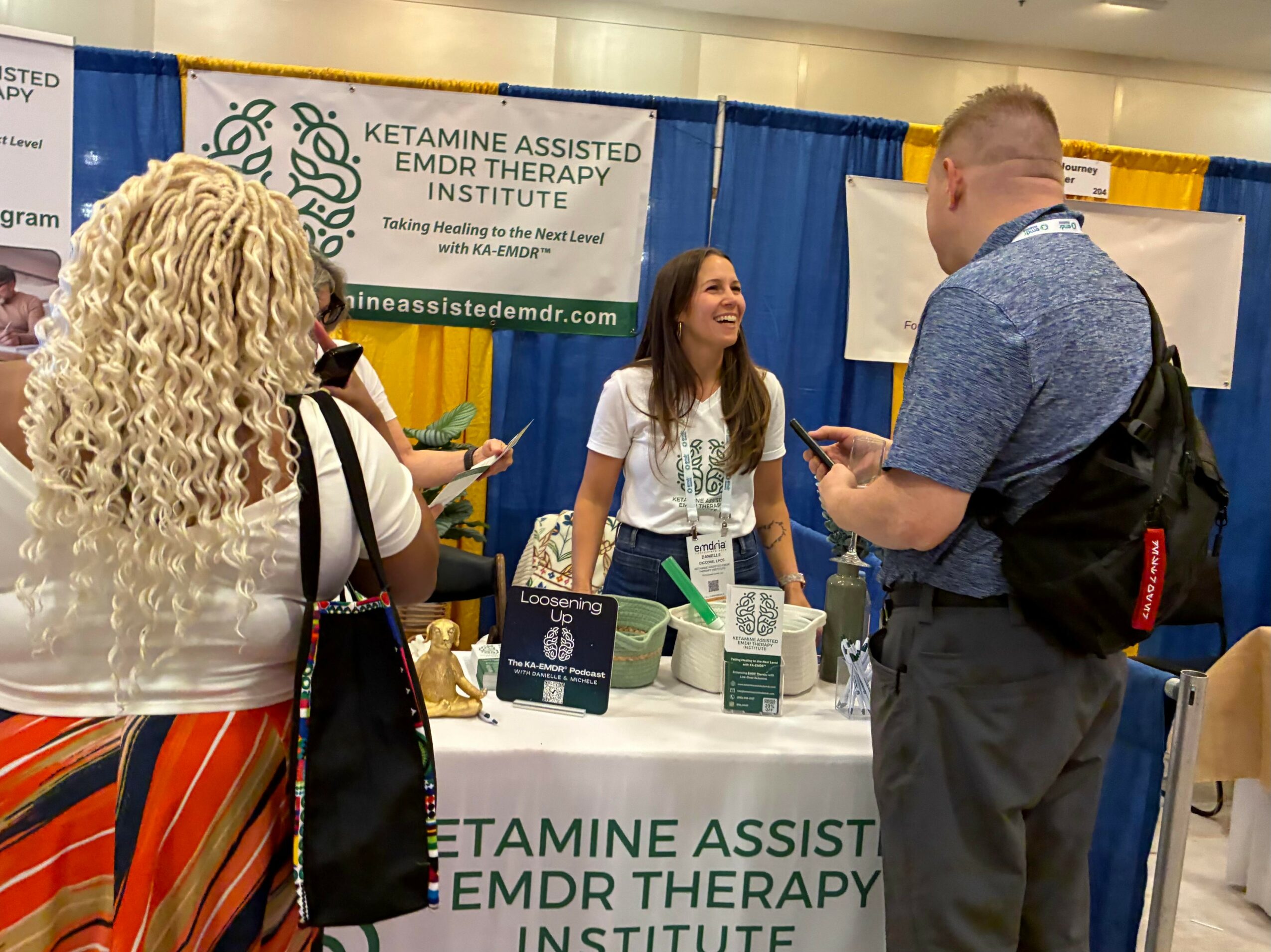Three people at a booth with a banner for Ketamine Assisted EMDR Therapy Institute, engaging in conversation, with a woman smiling.