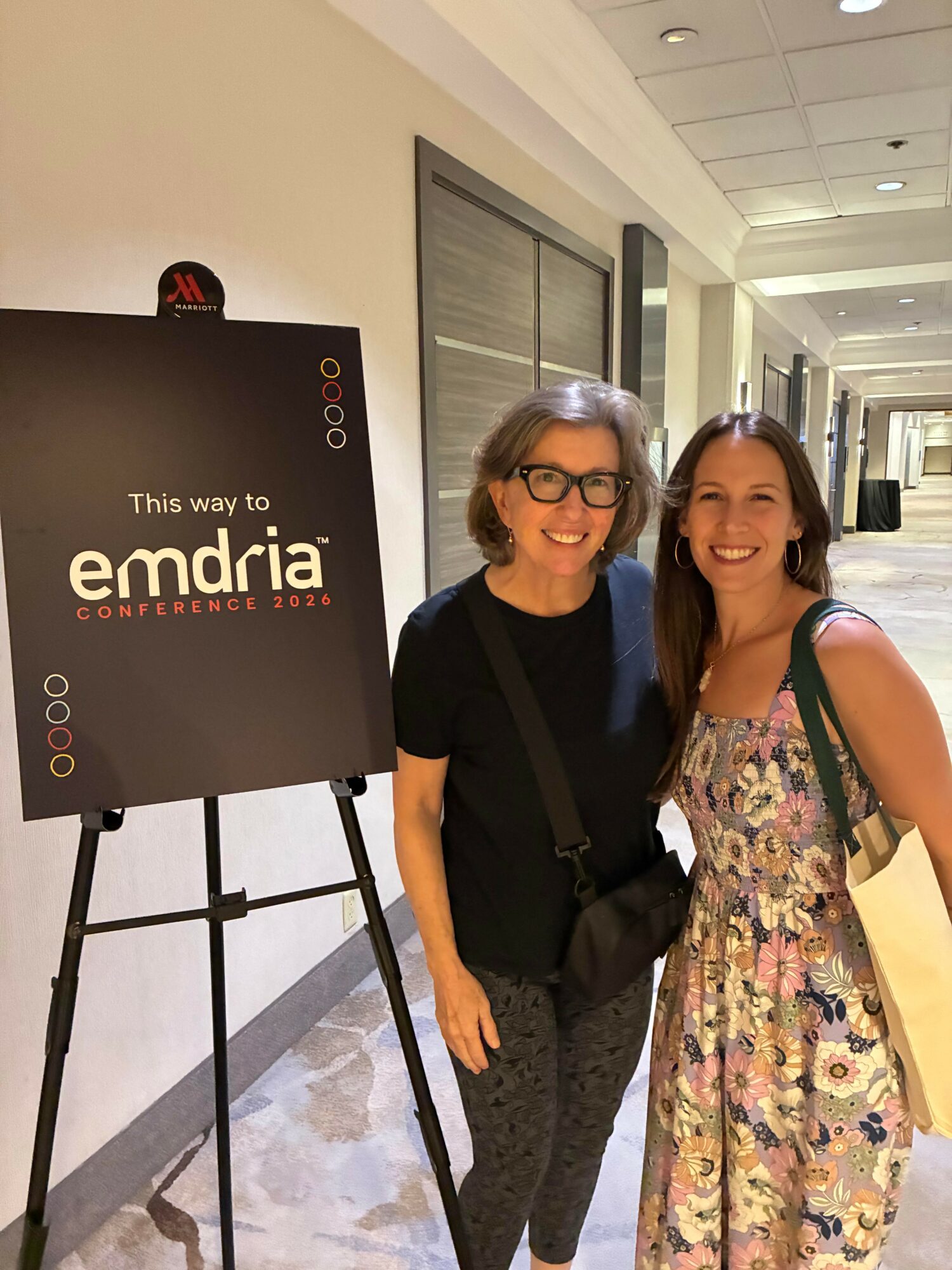 Two women standing indoors next to a sign for emdria conference 2023, smiling at the camera.