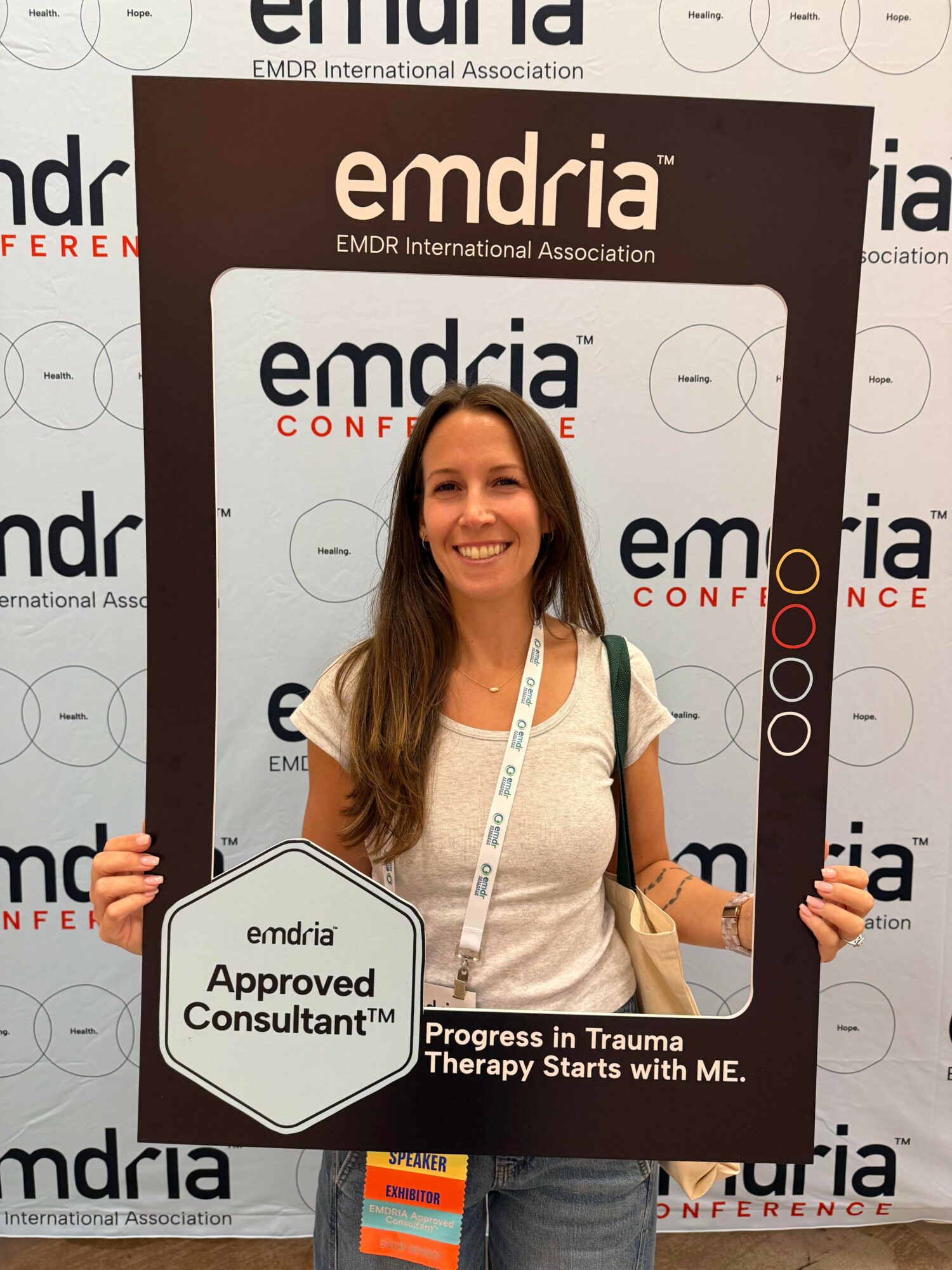 Woman smiling holding a frame at conference, background with 'emdria' logos, badge around neck, black and white frame.