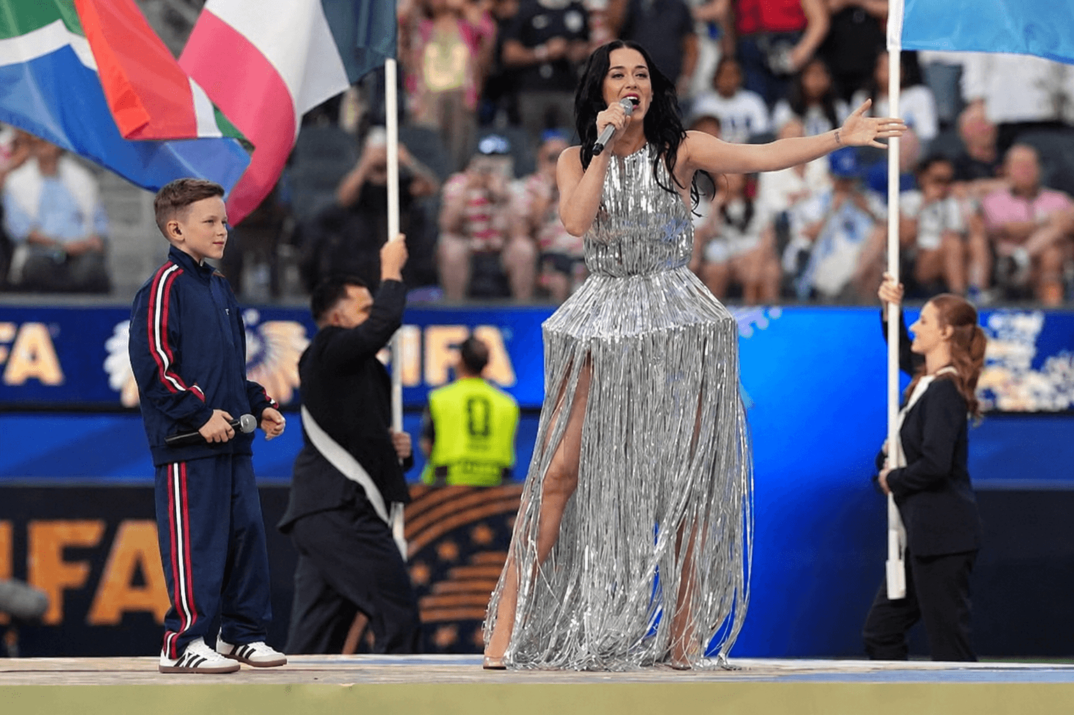Woman in a silver, fringed dress speaking into a microphone on stage with flags and audience in background.