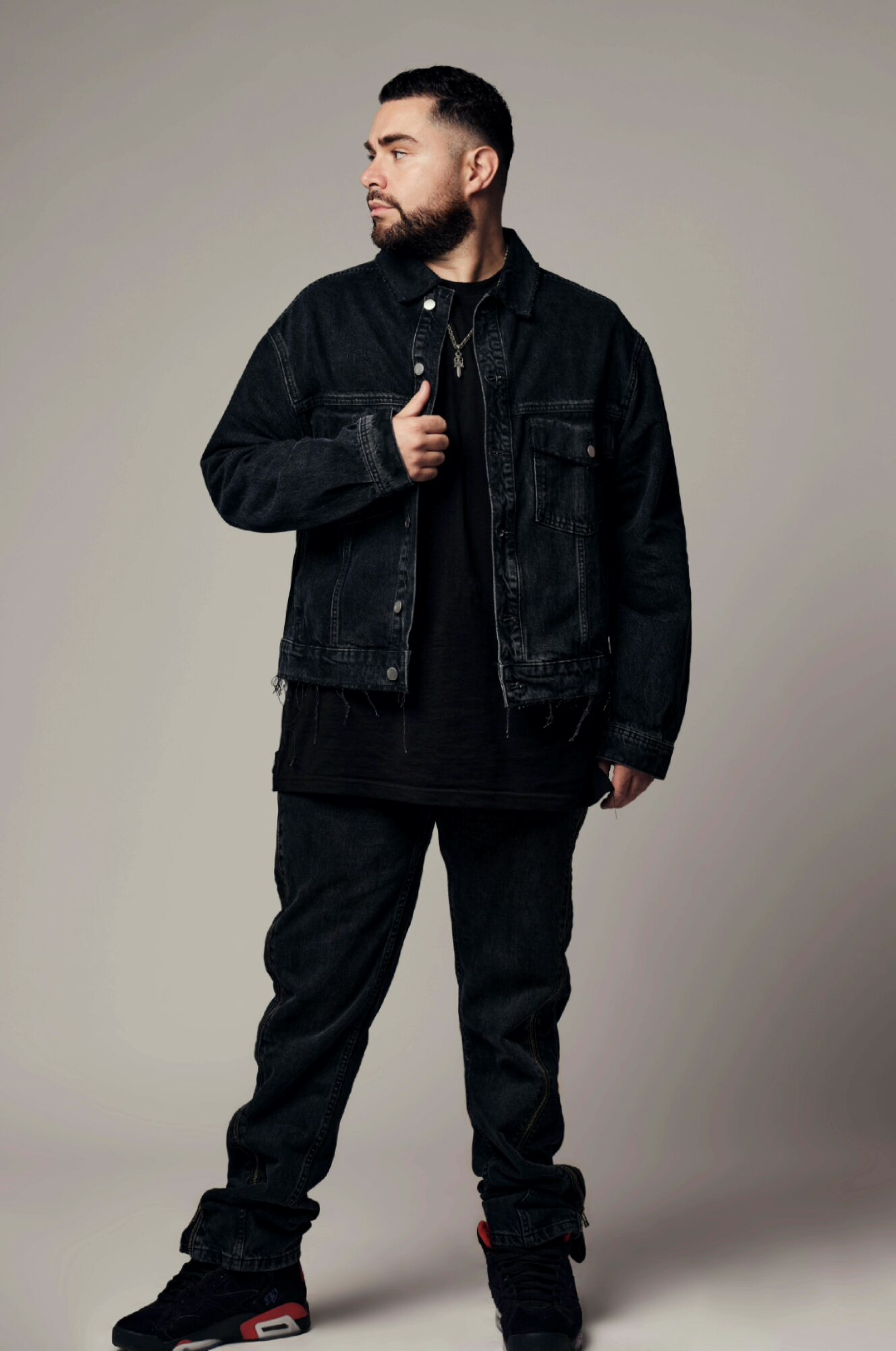 Man in black denim jacket and pants standing sideways, looking to the left, with a neutral background.