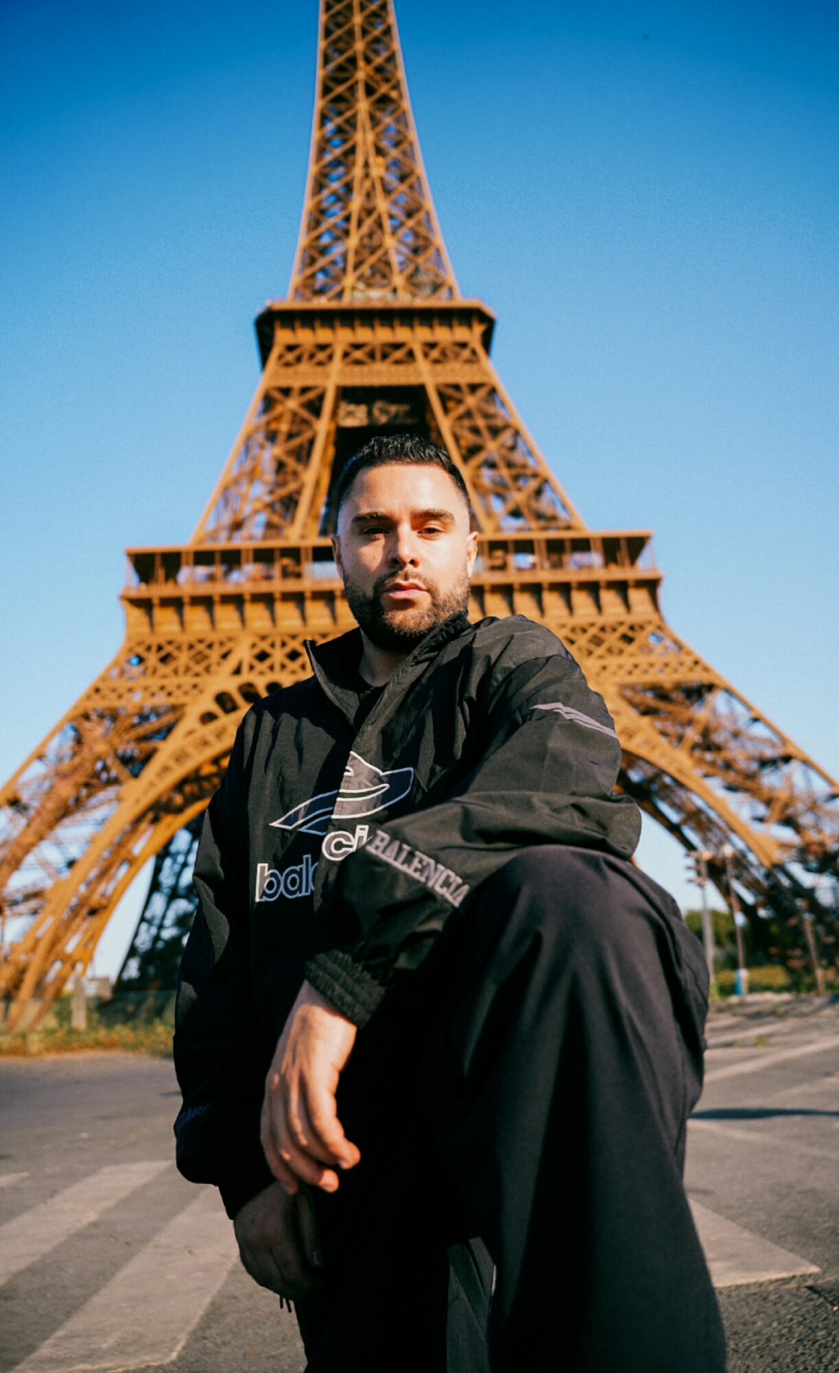Man with beard in black jacket sitting in front of Eiffel Tower during daytime.