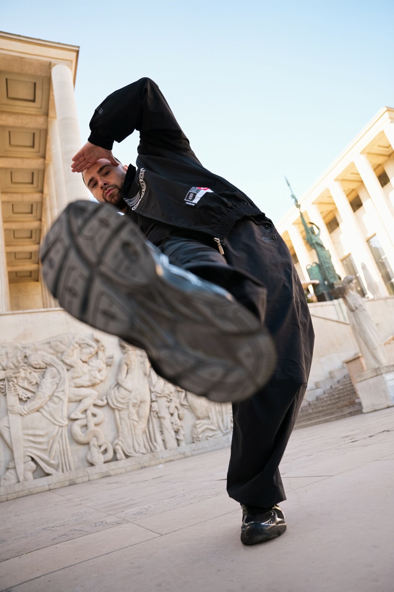 Person performing a high kick outdoors, with buildings in the background, wearing dark clothing and sneakers.