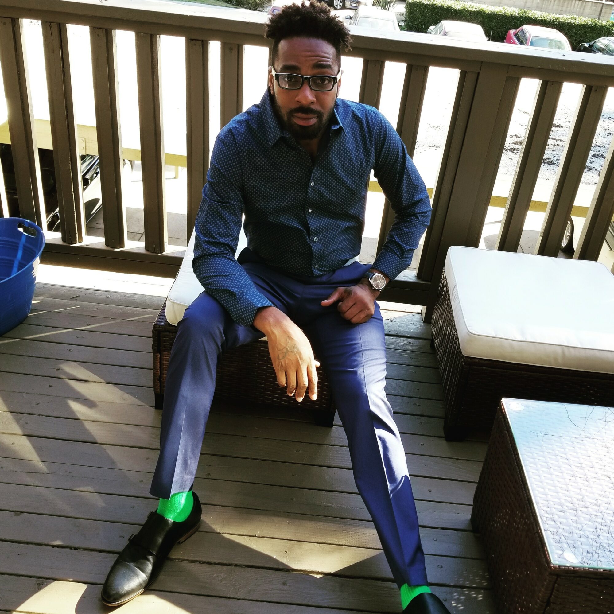 Man sitting on a wooden deck with a railing, wearing glasses, a blue shirt, and dark pants, with a green sock visible.