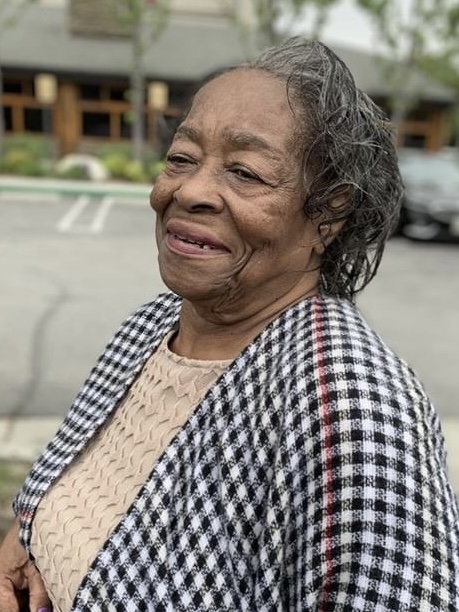 Elderly woman with gray hair smiling outdoors, wearing a checkered jacket and beige top.