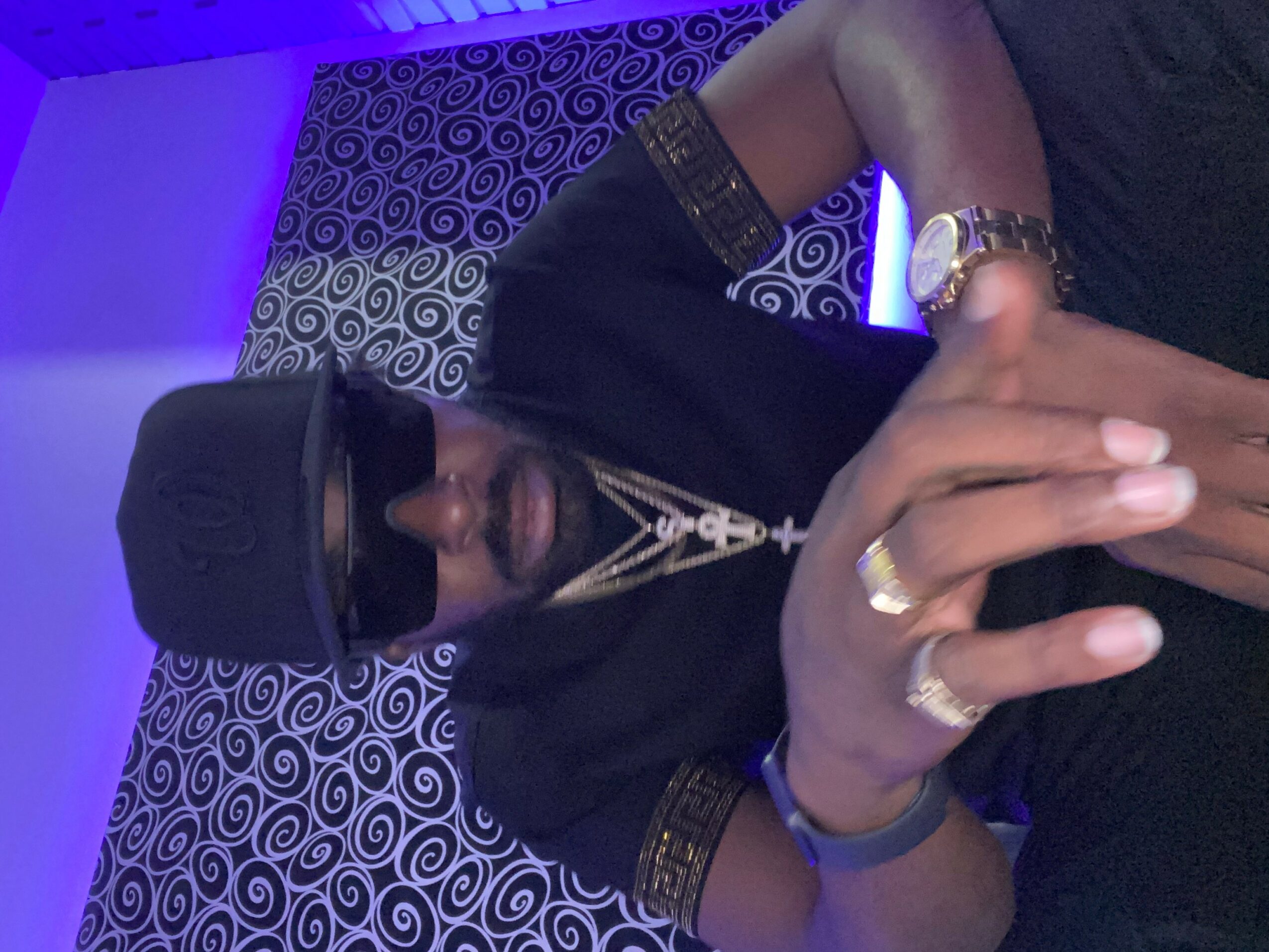 Person wearing a black cap, sunglasses, and jewelry, sitting on a patterned surface with blue lighting.