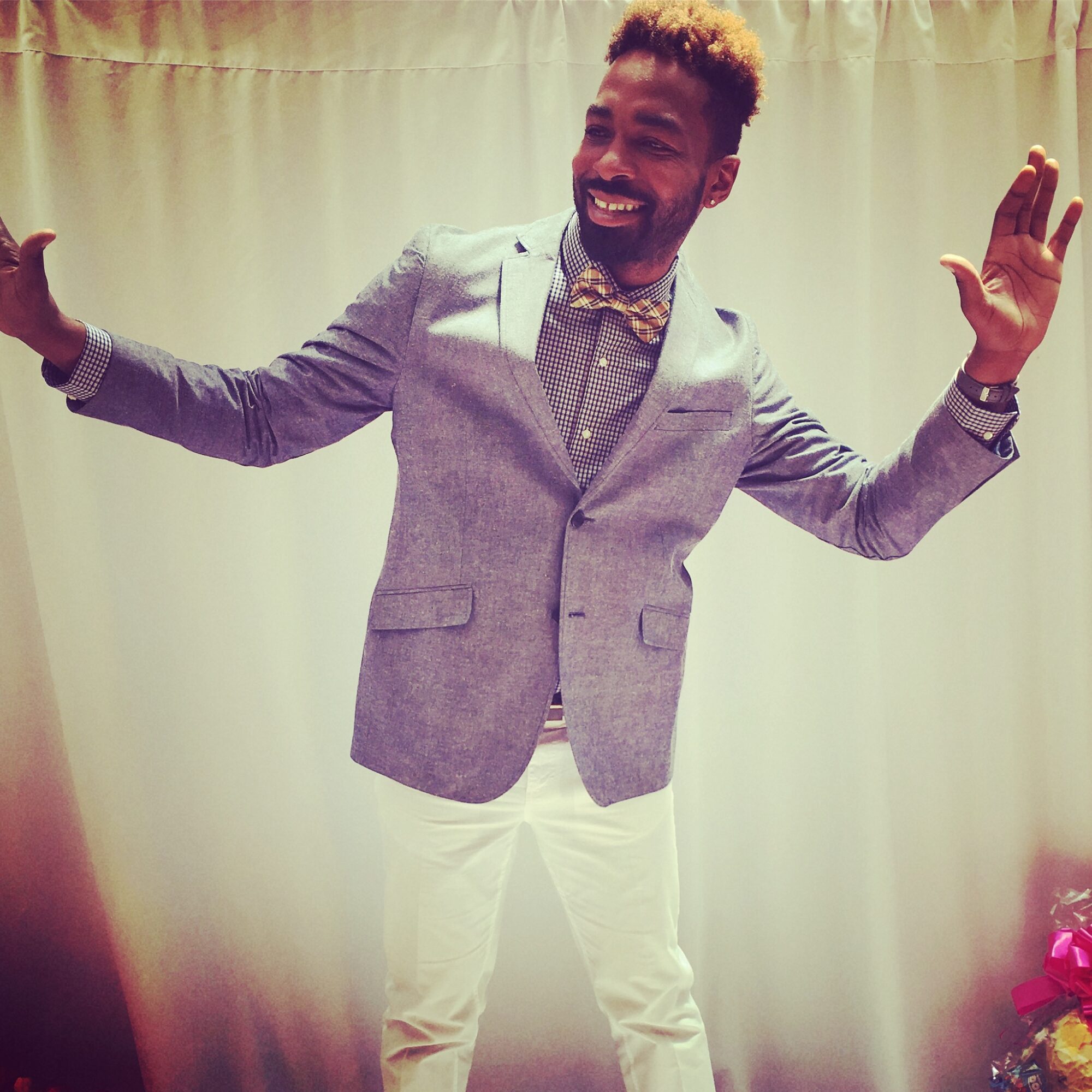 Person smiling with arms raised, wearing a gray blazer, bow tie, and white pants, standing in front of a curtain.