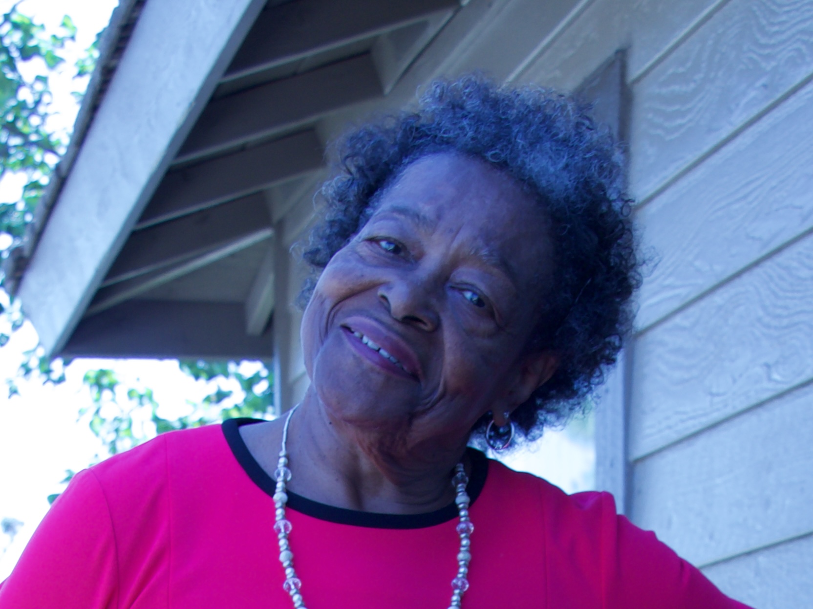 Older woman with curly hair wearing a pink top and pearl necklace, smiling outdoors near a house.