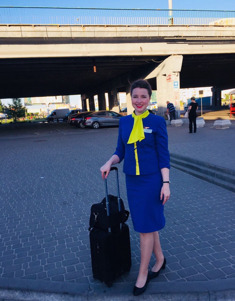 Woman in blue suit with yellow scarf standing outdoors with luggage under a bridge, smiling.