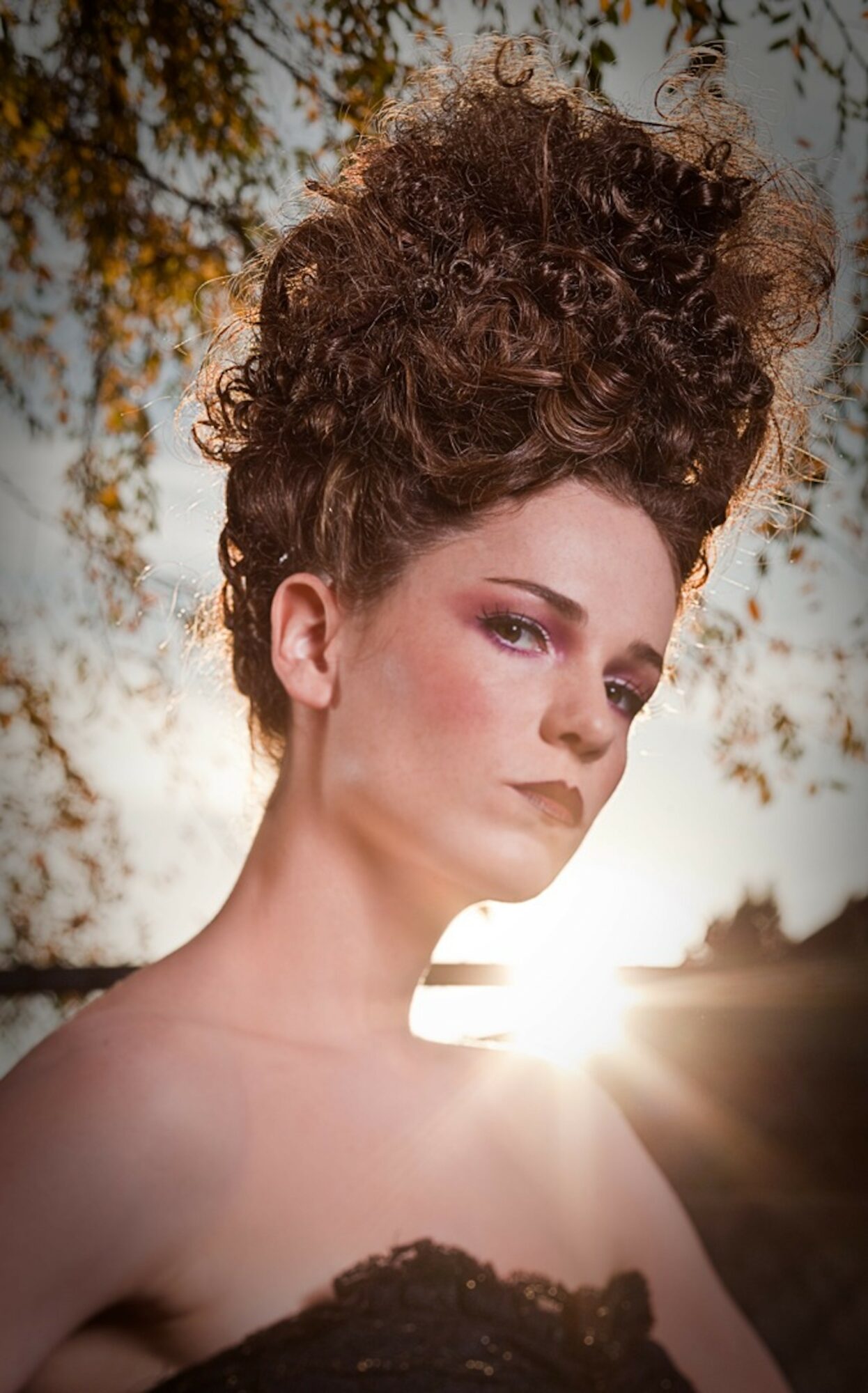 Portrait of a woman with curly hair and makeup, outdoors with sunlight behind her, trees in the background.