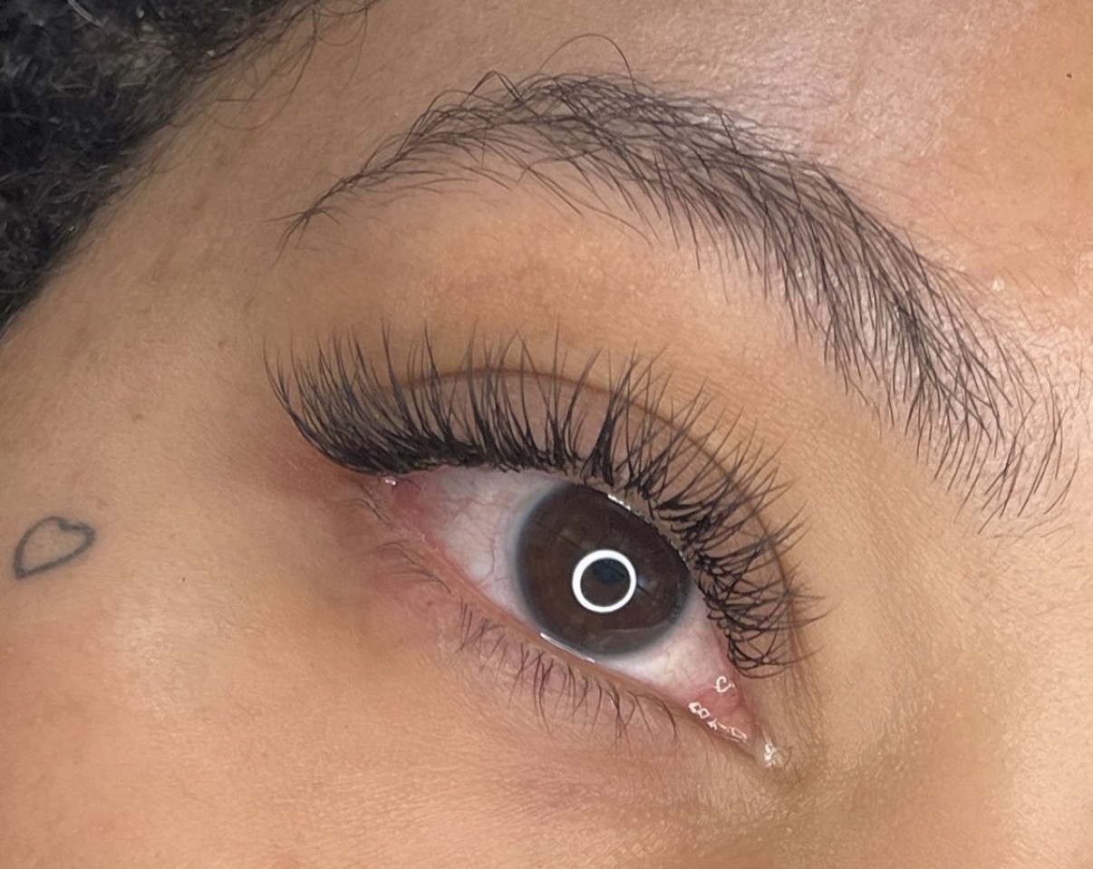 Close-up of a person's eye with long eyelashes and a small heart tattoo near the eyebrow.