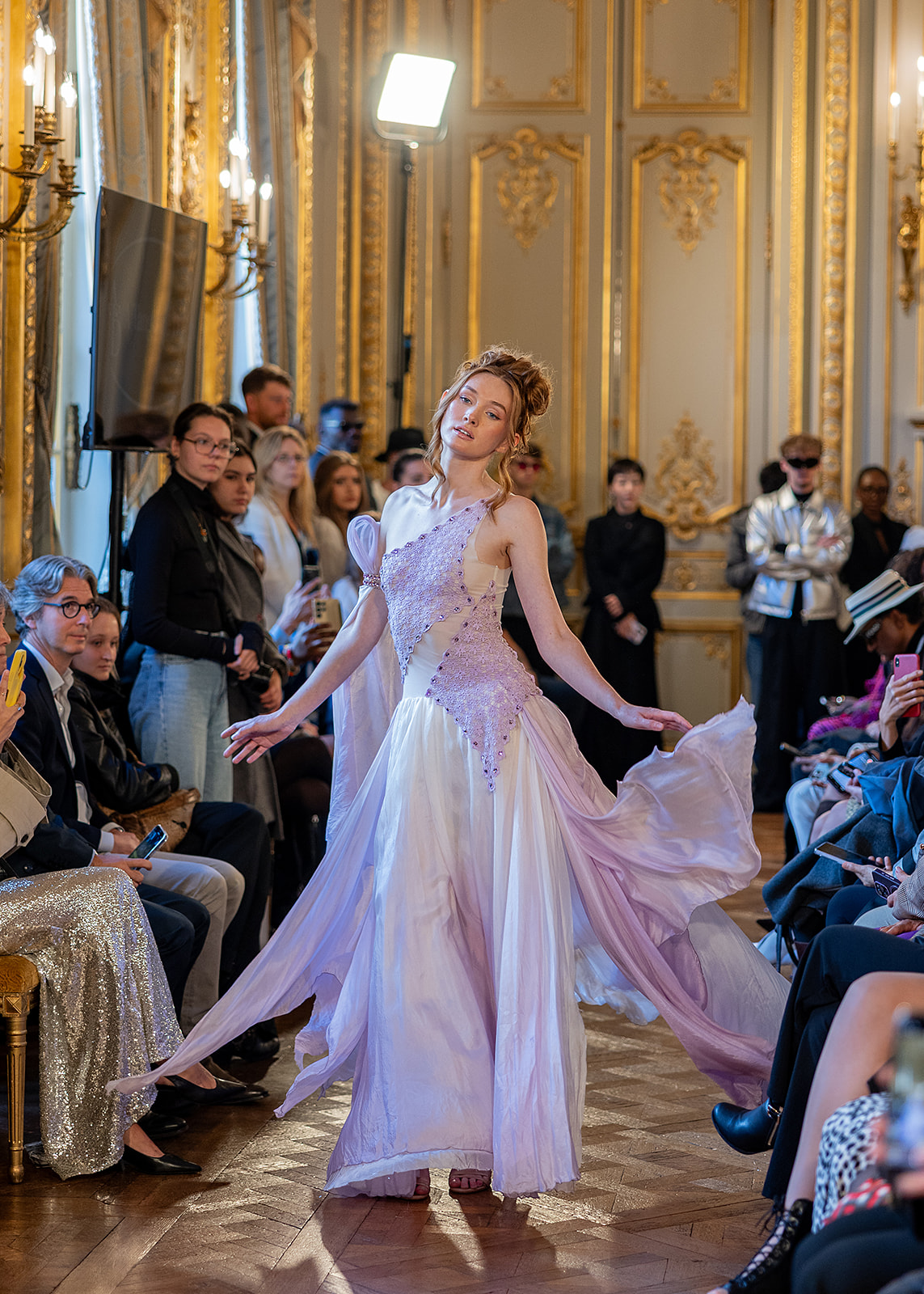 A woman in a flowing white dress dances in a grand, ornate room with an audience watching.