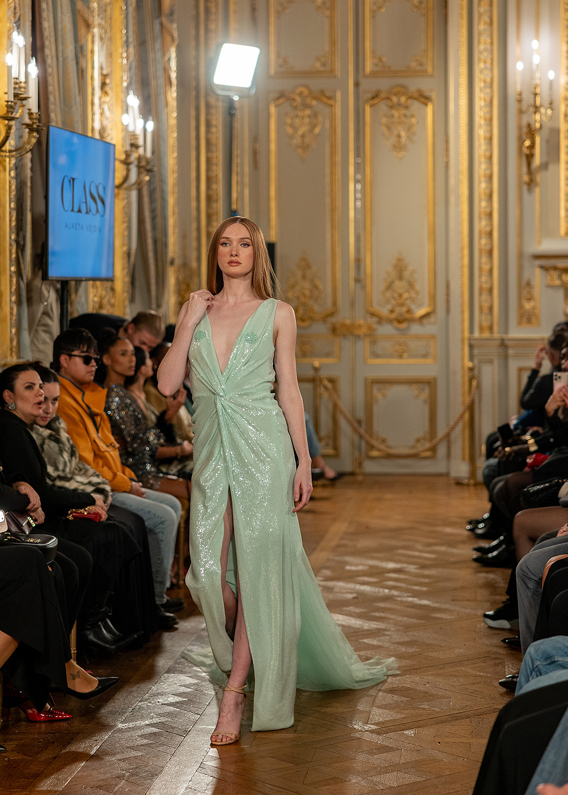 Model walking down runway in elegant green gown with slit, audience seated on both sides, ornate gold decor in background.