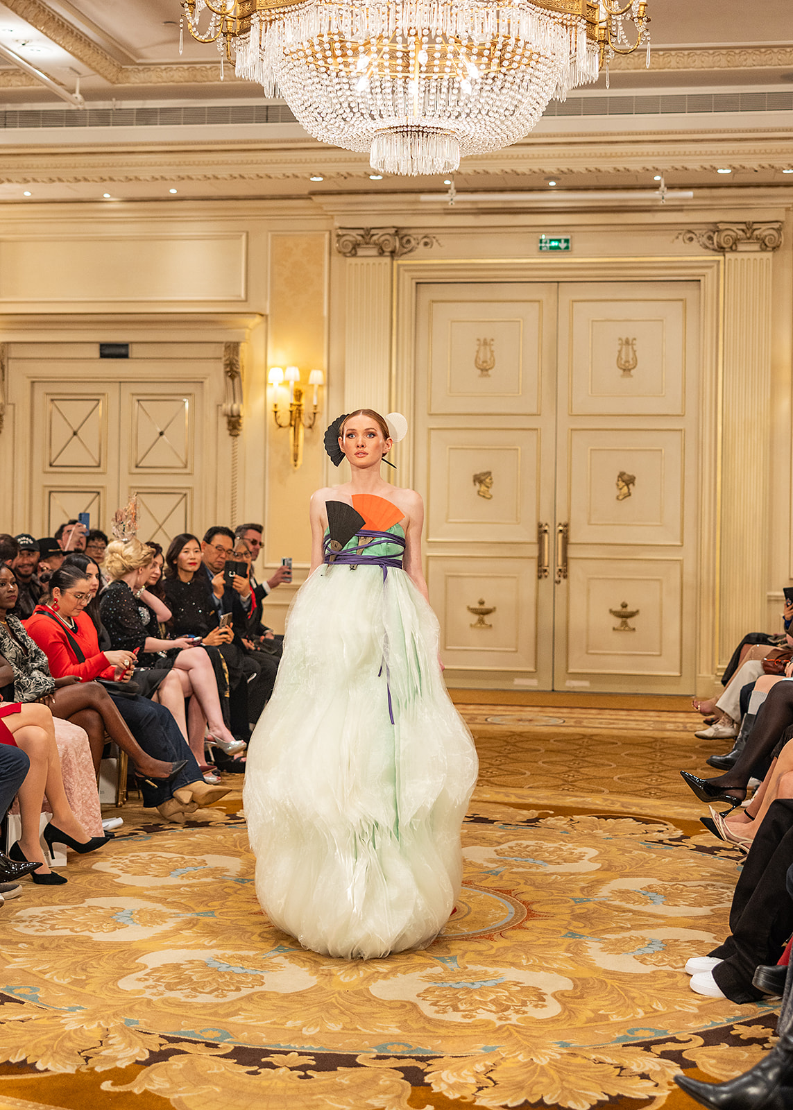 Woman in a strapless gown walking on a runway in a grand room with chandeliers and ornate doors, audience seated on both sides.