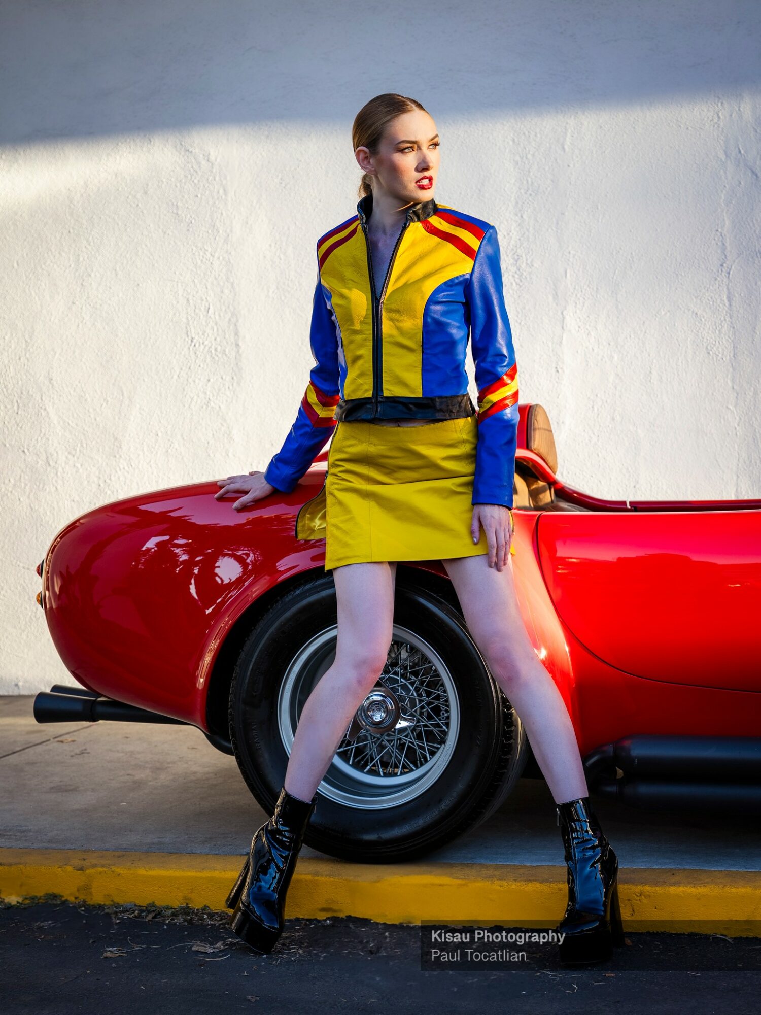 Woman in colorful jacket and yellow skirt stands next to a red vintage car against a white wall.