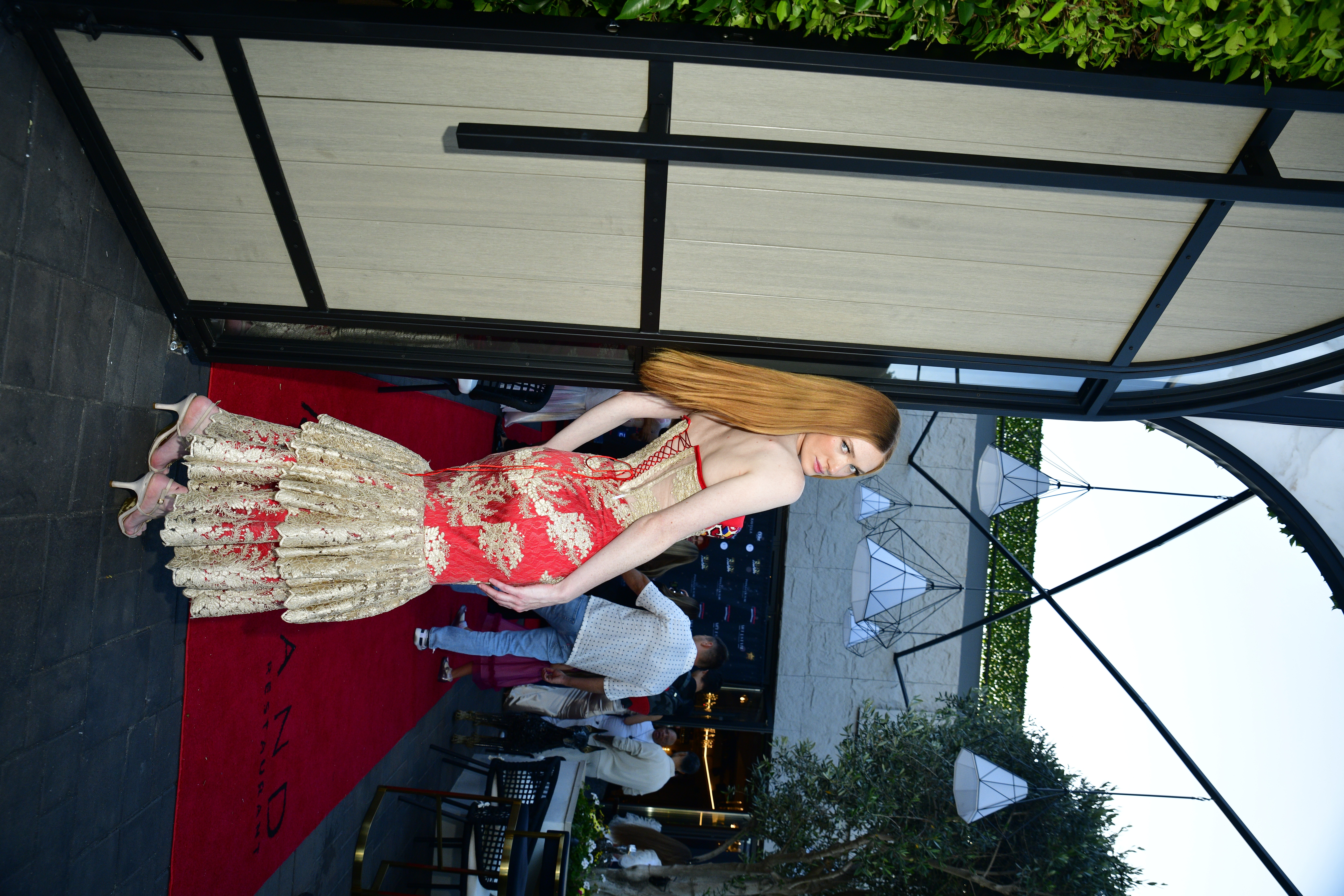 Person in a red and gold dress standing on a red carpet outside near a white fence, with a tent and trees in background.