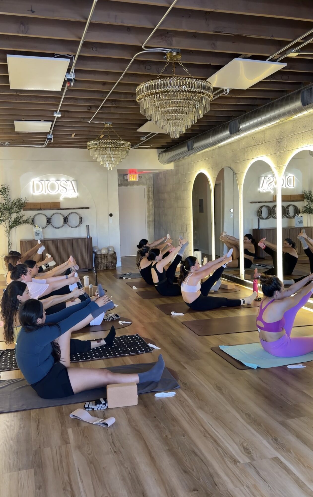 Group of people practicing yoga in a studio with chandeliers and arched windows, sitting on mats with legs raised.