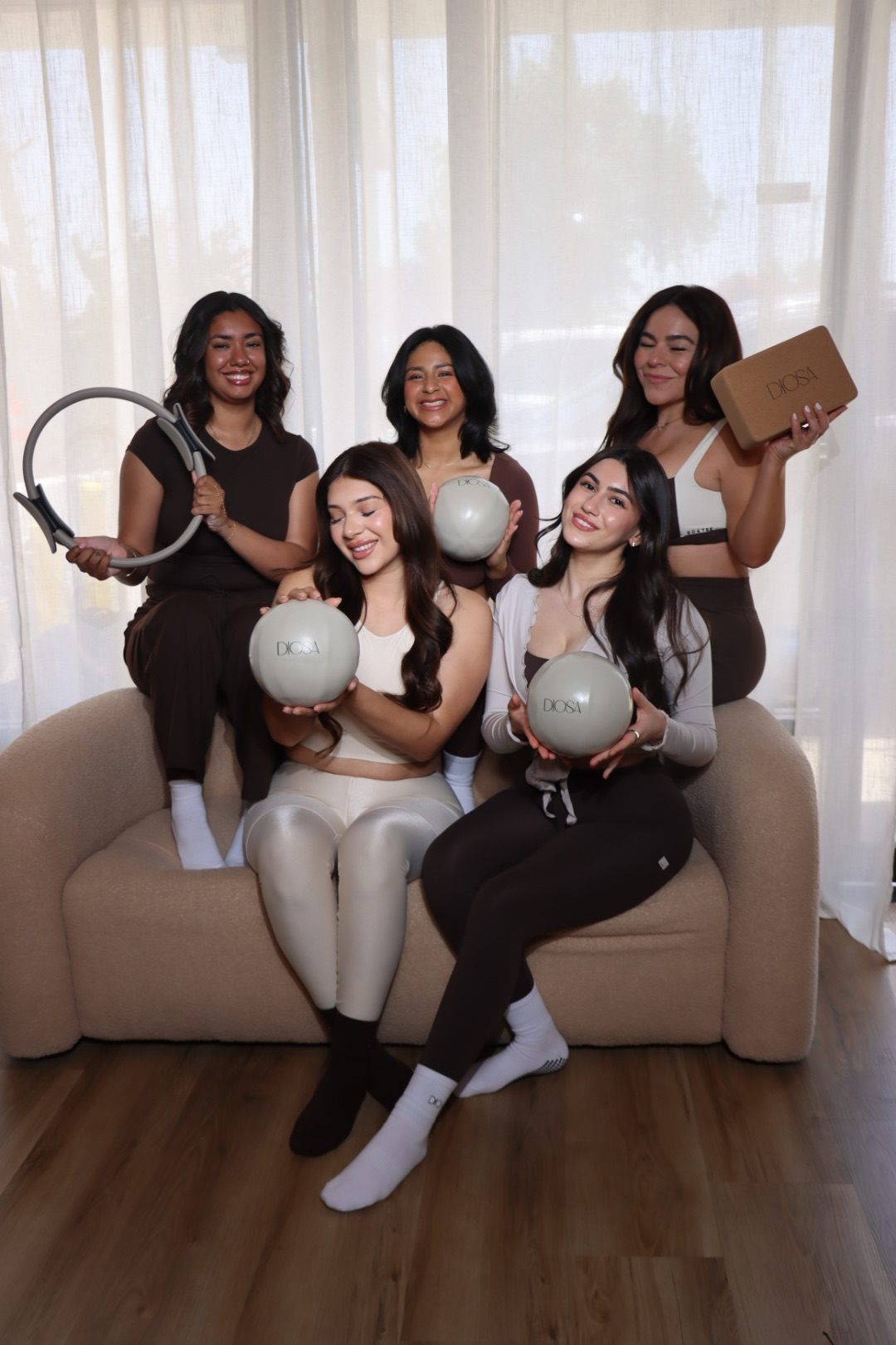 Five women sitting on a sofa holding various objects, with a curtain background and wooden floor.