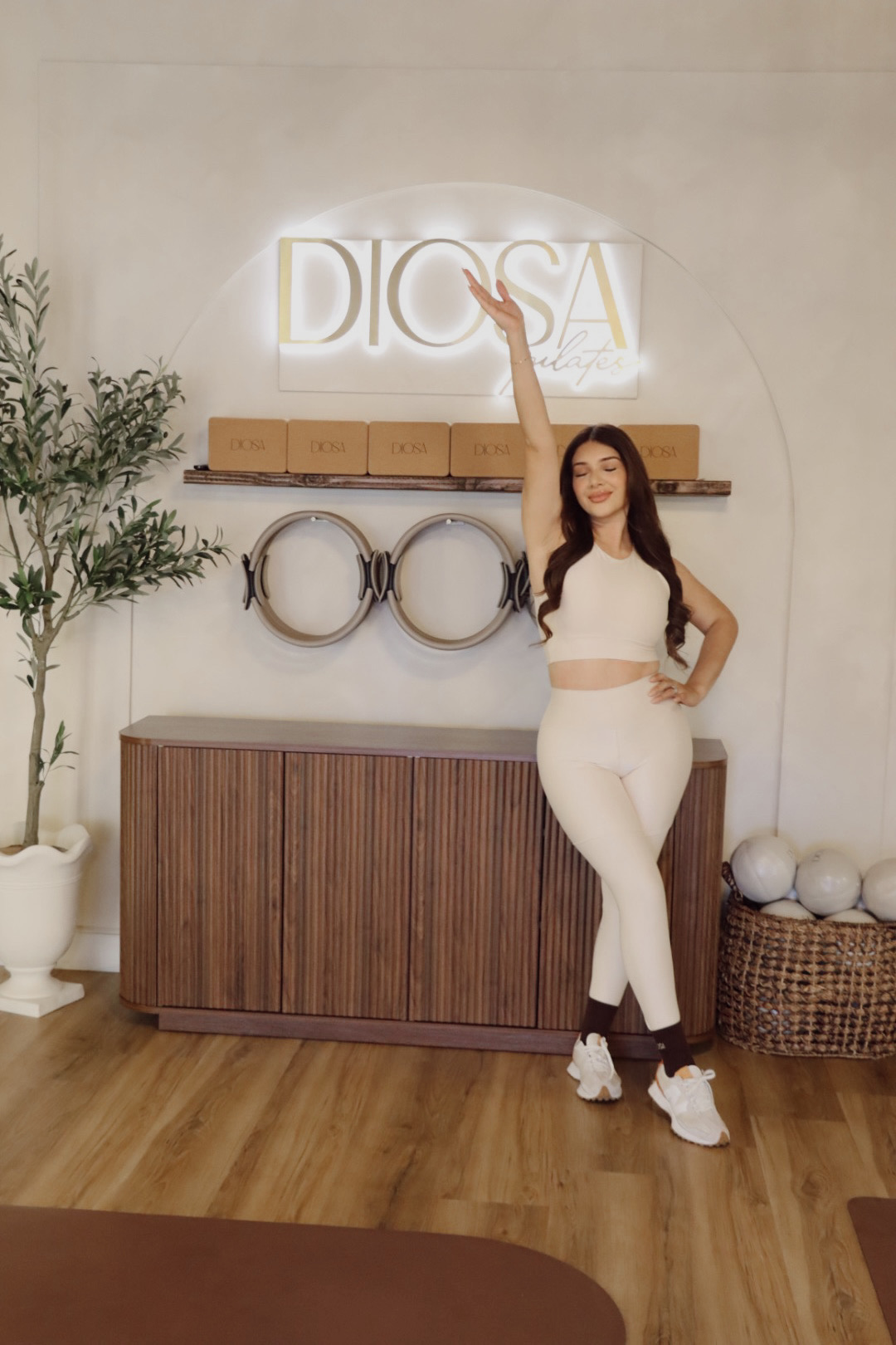 Woman in white outfit standing in front of a wall with a neon sign that says 'DIOSA', with decor including a potted plant and a wooden cabinet.