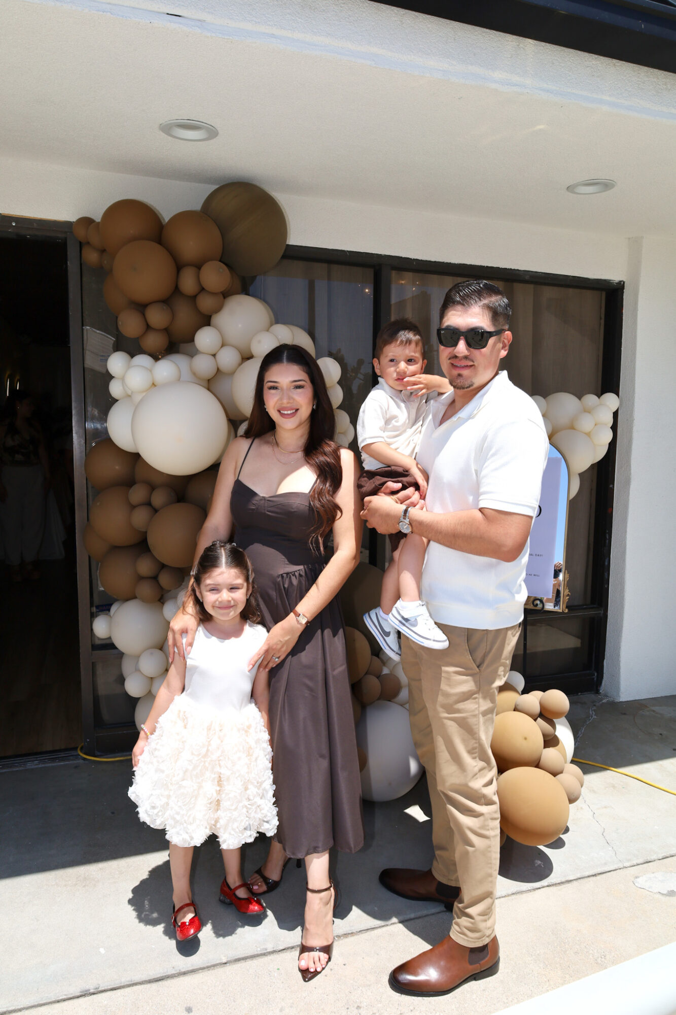 Family of four standing outside a building with balloon decorations, smiling, with a woman in a brown dress, man in white shirt, and two children.