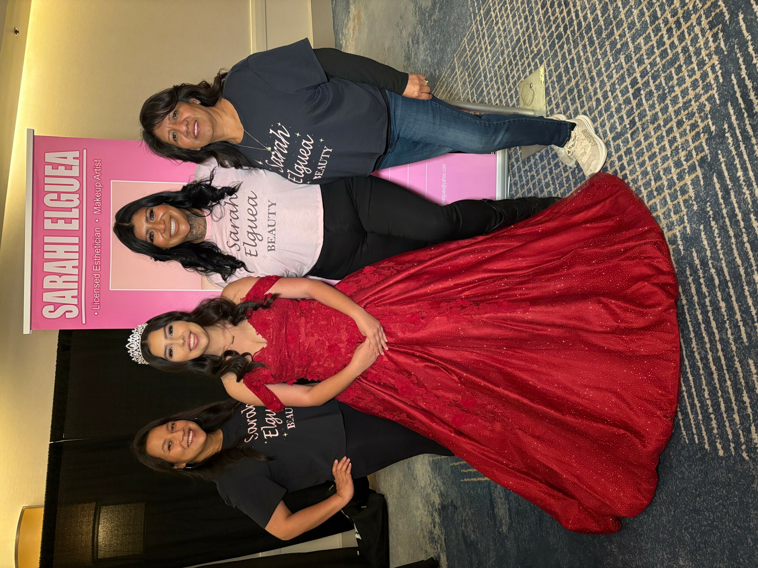 Four women posing together, one in a red gown, three in black tops, standing in front of a pink sign with text.