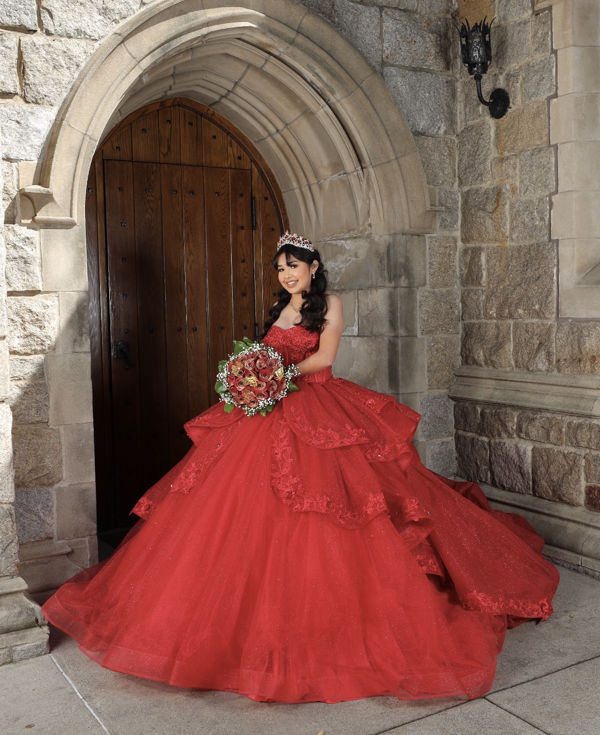 Young woman in a red gown holding a bouquet, standing in front of a stone archway and wooden door.