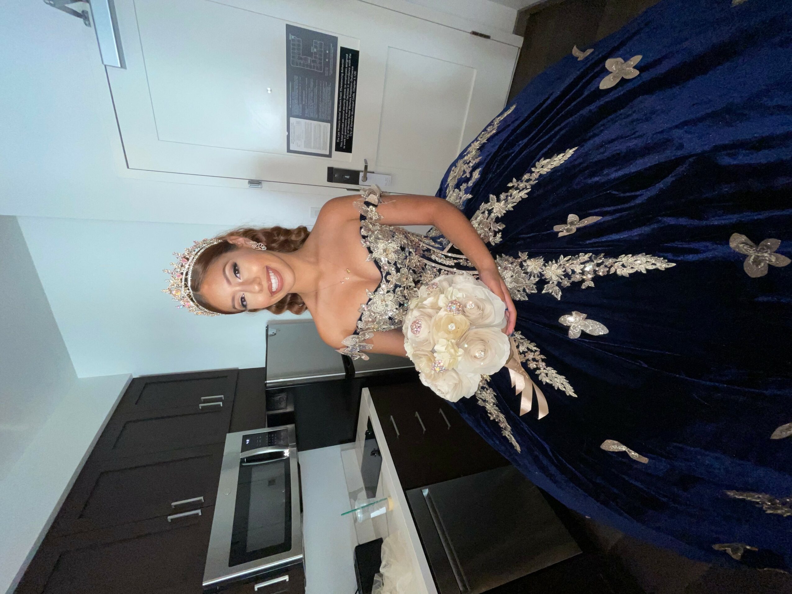 Woman in a blue gown holding a bouquet, standing in a kitchen, smiling, with a tiara and jewelry.