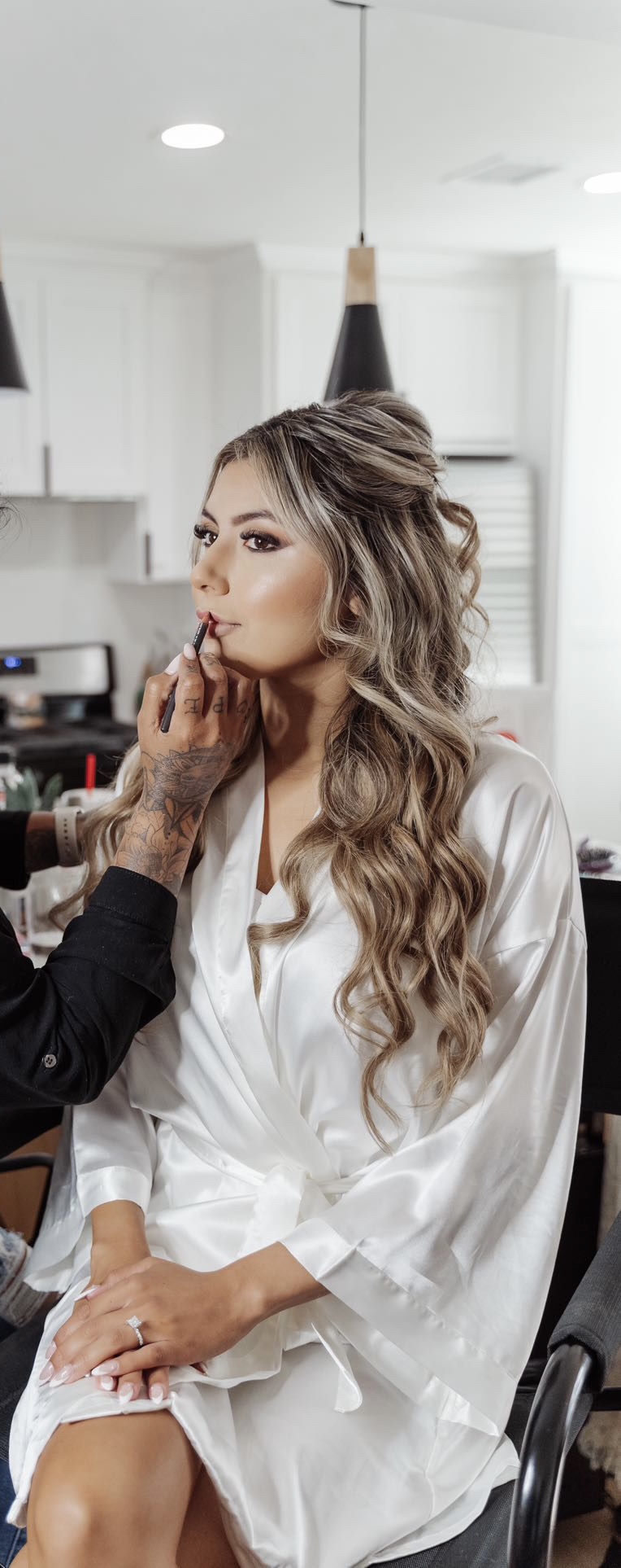 Woman with long wavy hair sitting in a chair, getting makeup applied, in a bright room with white walls.